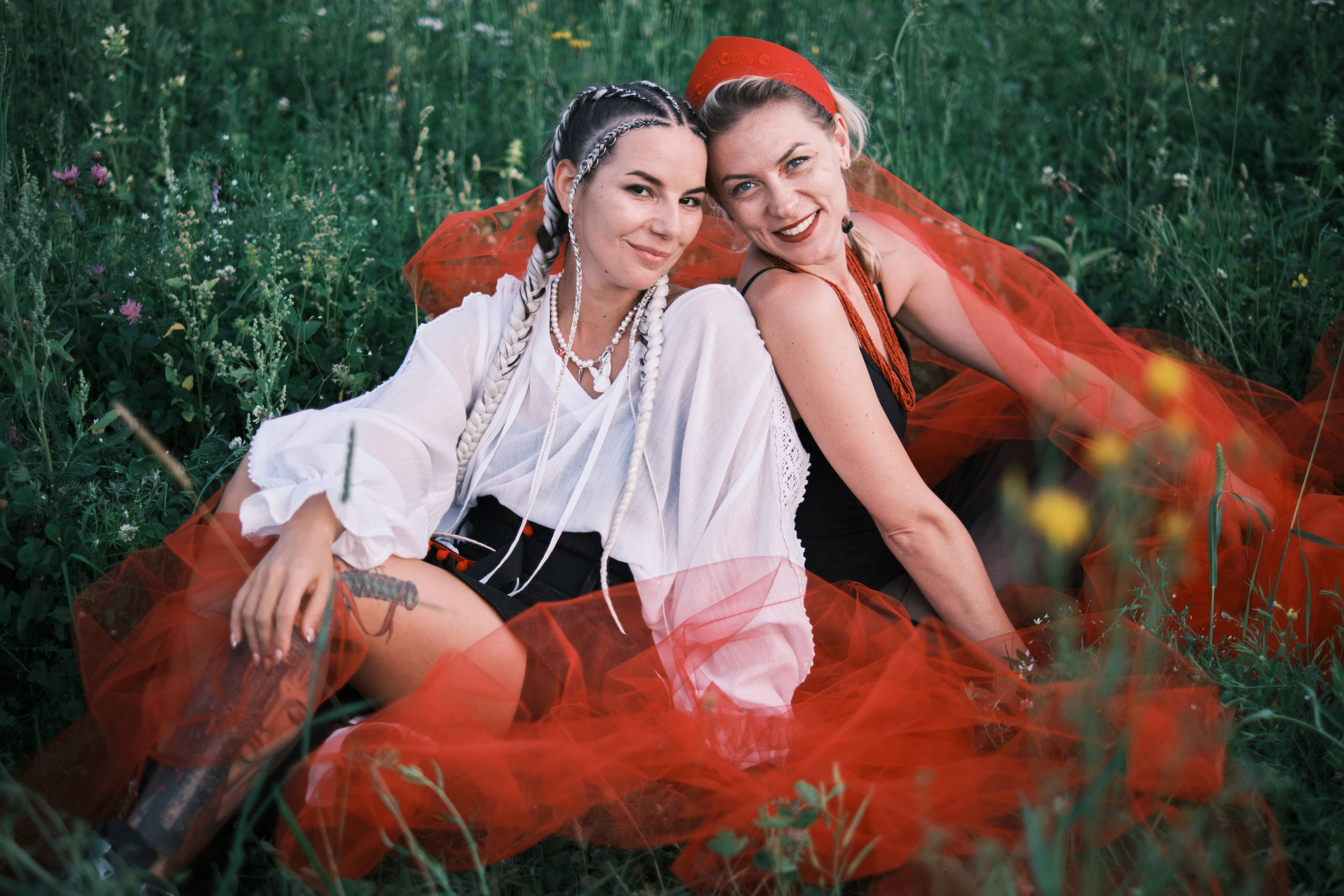 Две девушки сидят на природе, укрывшись широкой летящей тканью красного цвета.