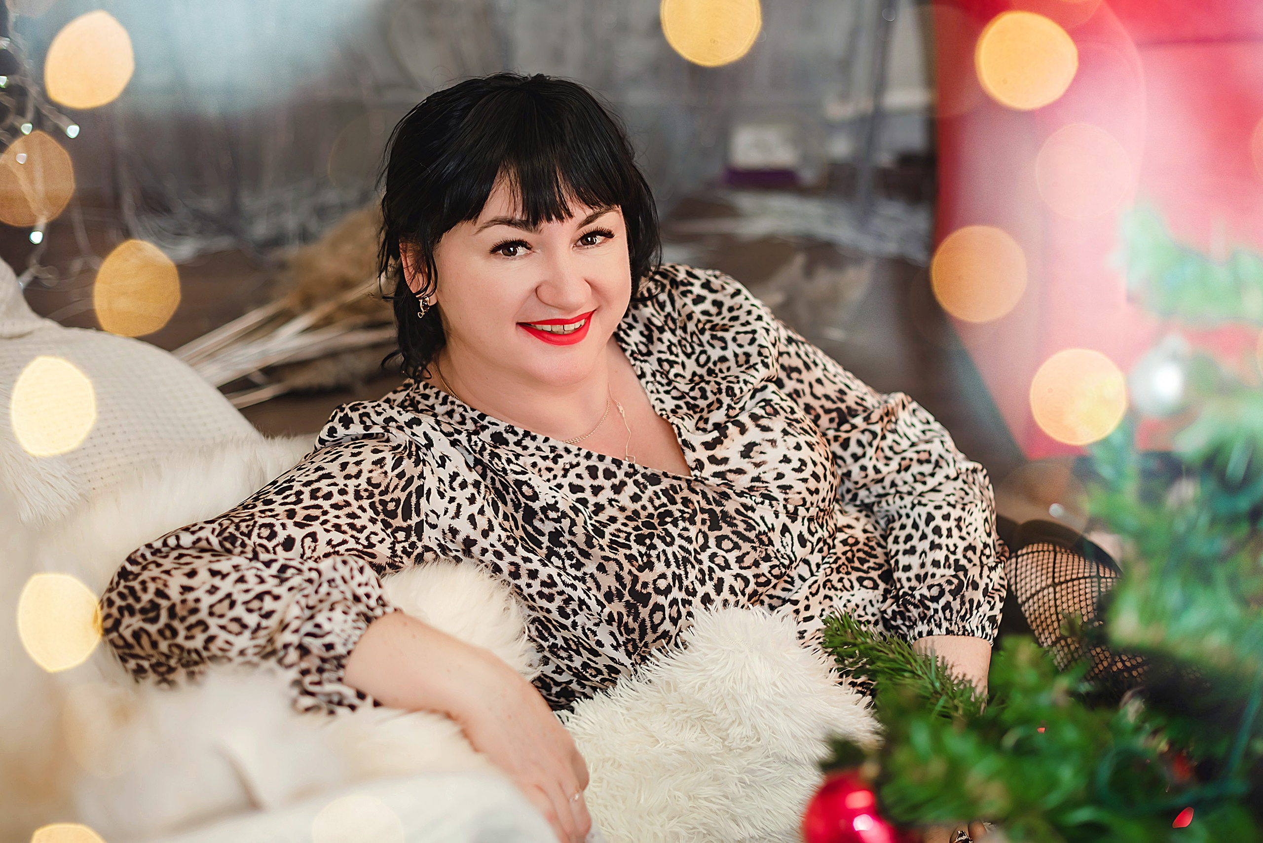 На фото  портрет девушки с подарком сидит около елки с огоньками на переднем плане.