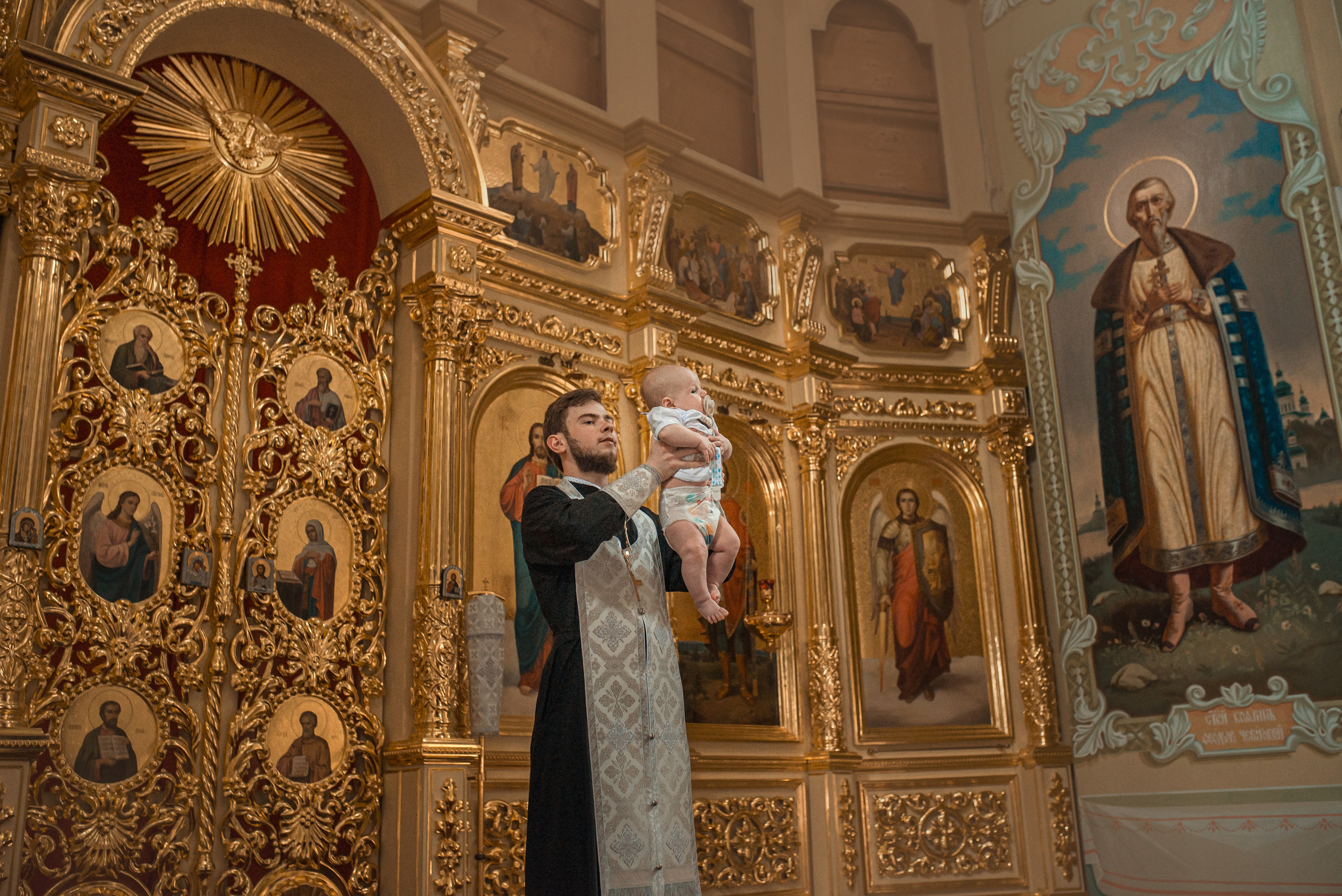 батюшка держит на руках ребенка в покровском соборе города саратова, крещение в саратове