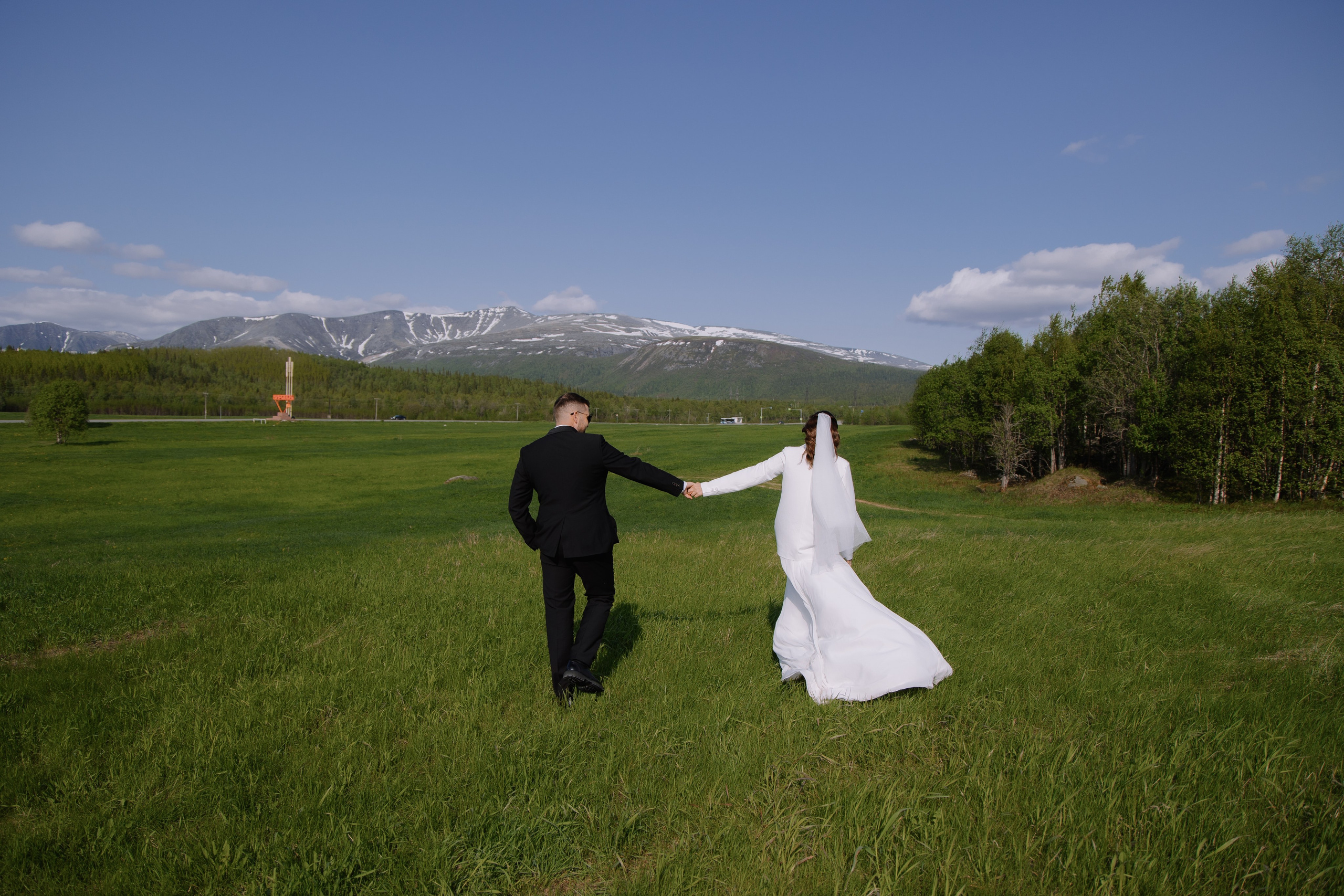 Жизнерадостная невеста идет босиком по мягкой траве, ведя за собой жениха.