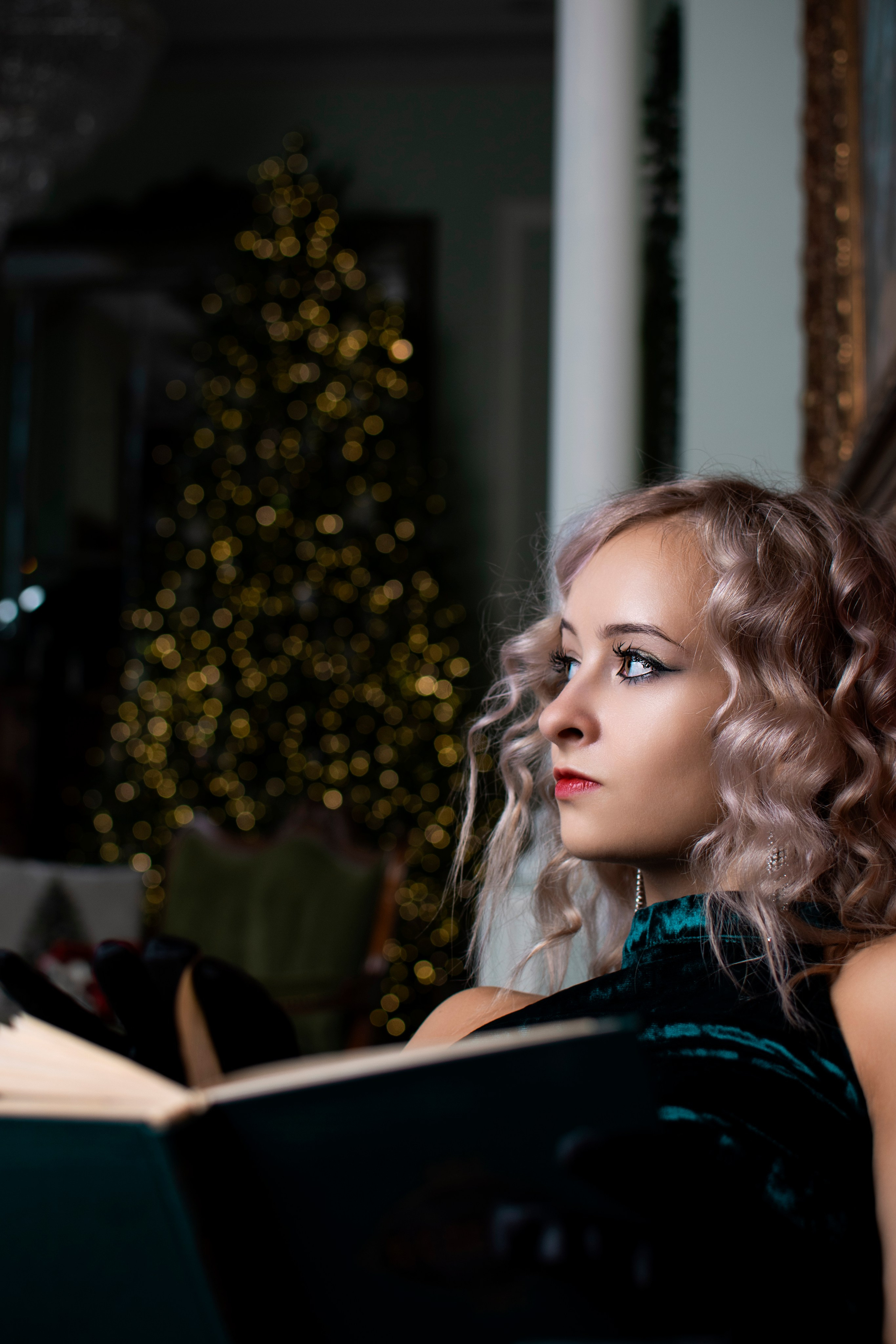Кудрявая светловолосая девушка читает книгу у Новогодней лки