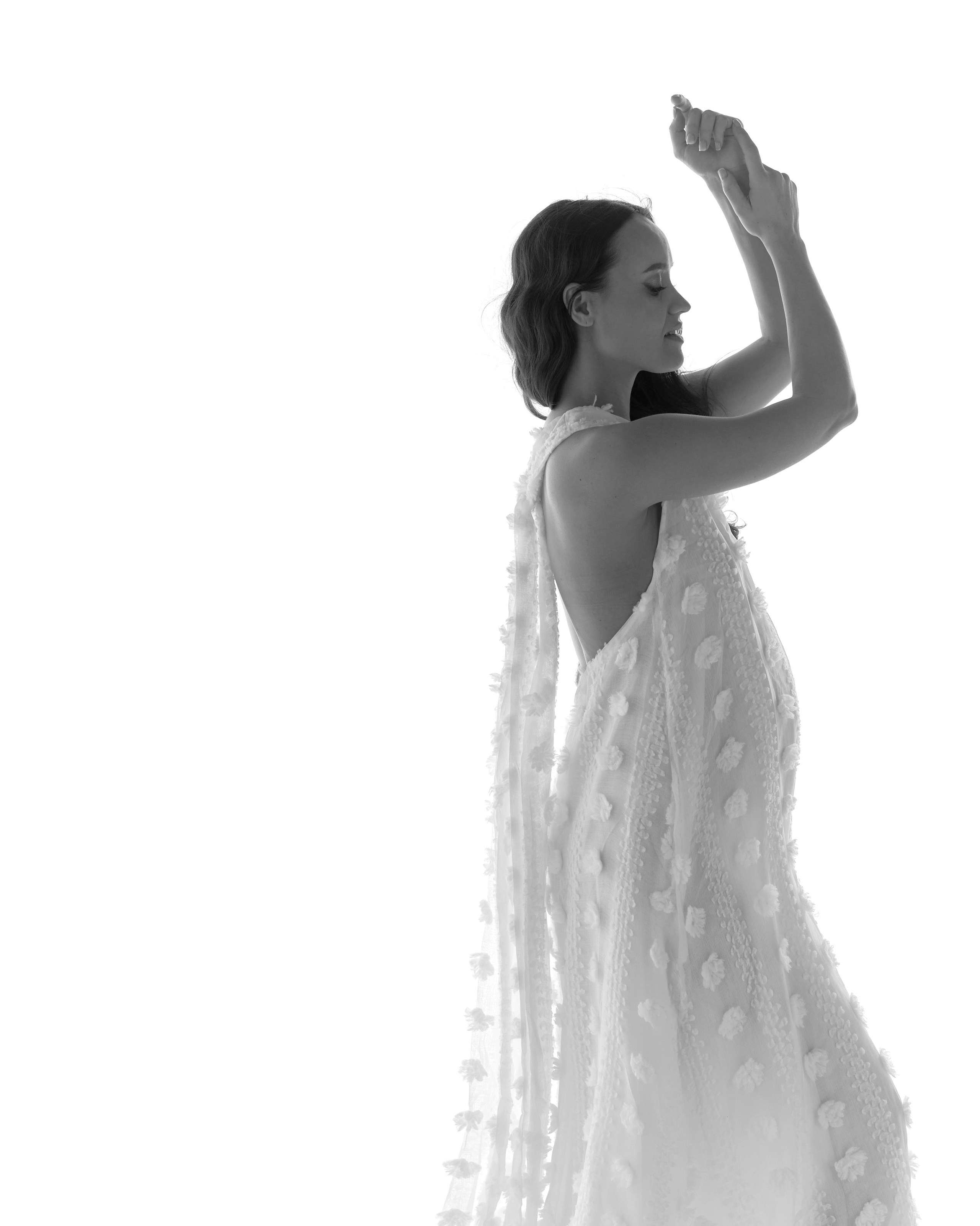 Черно-белый портрет беременной в изысканном белом платье с объемными цветочными аппликациями и открытой спиной. Она стоит в профиль, подняв руки, на белом фоне.