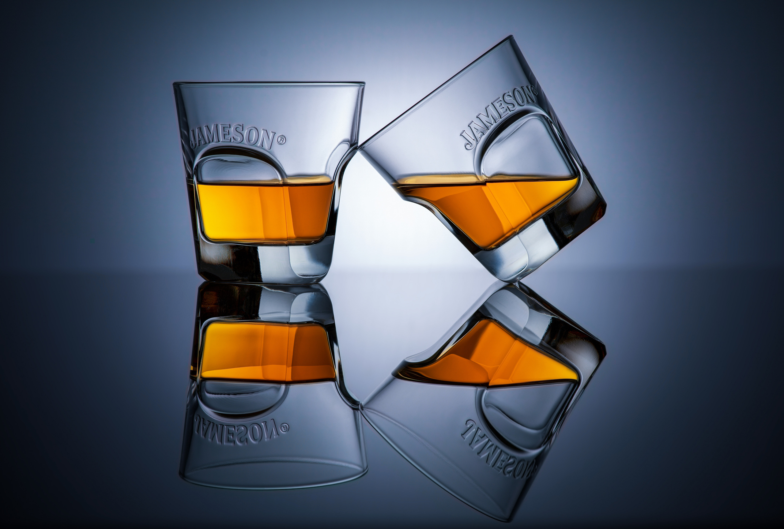  два  бокала Jamesson с виски один наклонен под 45 градусов и прислонён к другому на сером фоне с радиальным градиентом