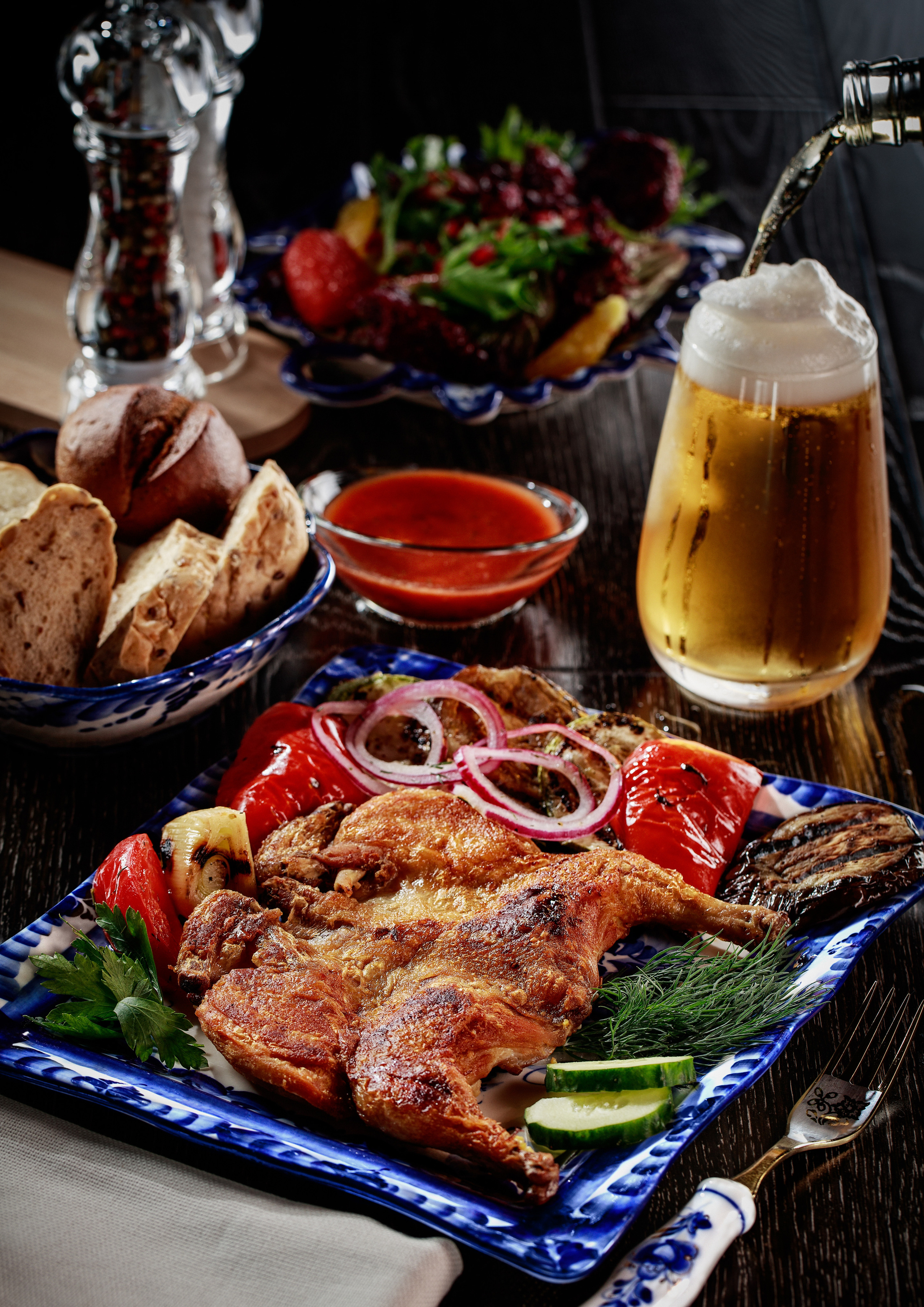 натюрморт -на переднем плане цыплёнок табака на гжелевской тарелке с гарниром из свежих огурцов и помидоров лука и зелени ,а на втором плане тарелка с хлебом розетка с соусом бокал с пивом которое наливается в него из бутылки,и на заднем плане мельницы с солью и перцем на разделочной доске и салат из печеной свёклы с каймаком с кедровыми орехами