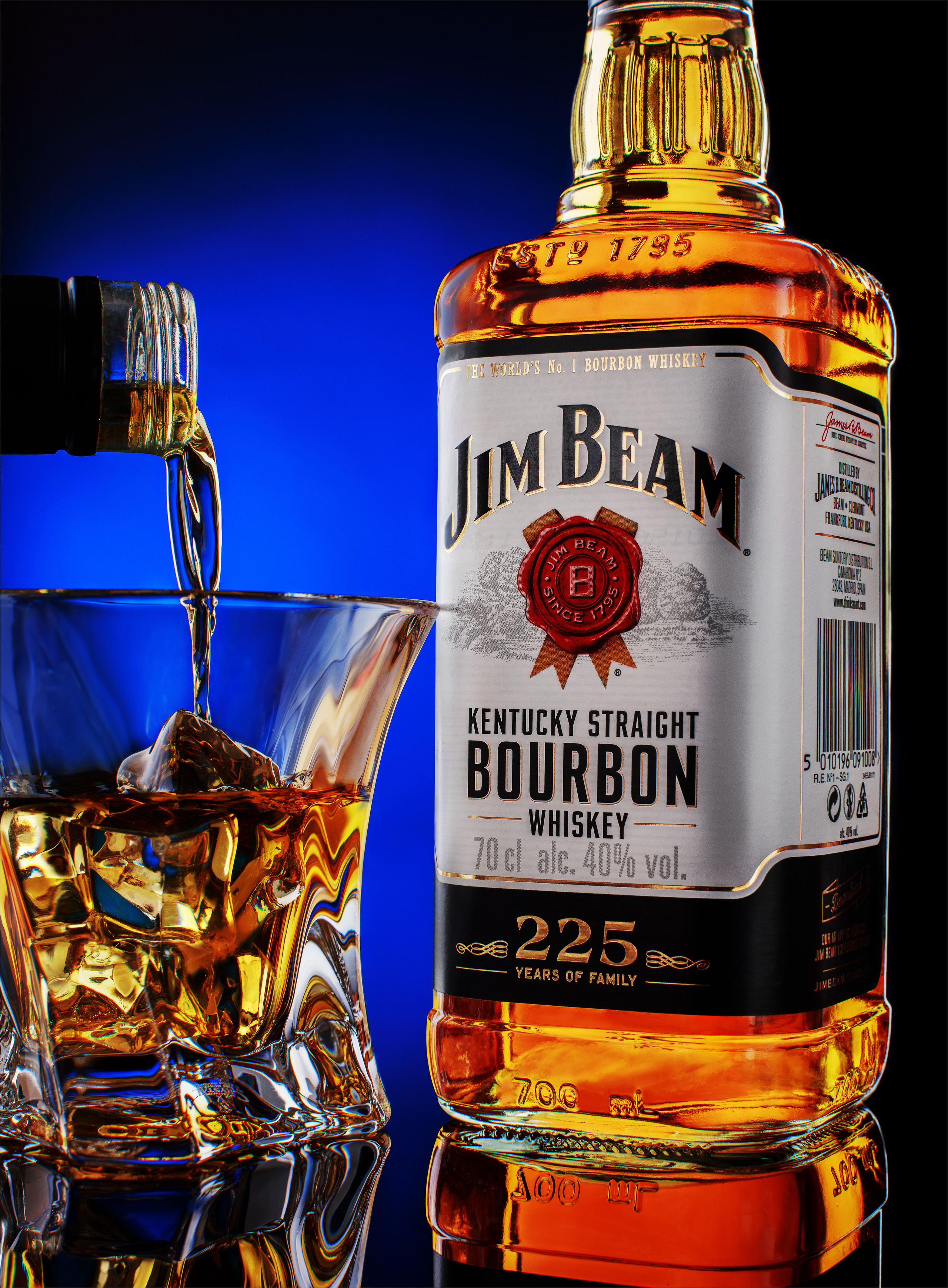 бутылка  бурбона JimBeam и стакан со льдом и бурбоном в который наливают напиток на фоне синего градиентного пятна