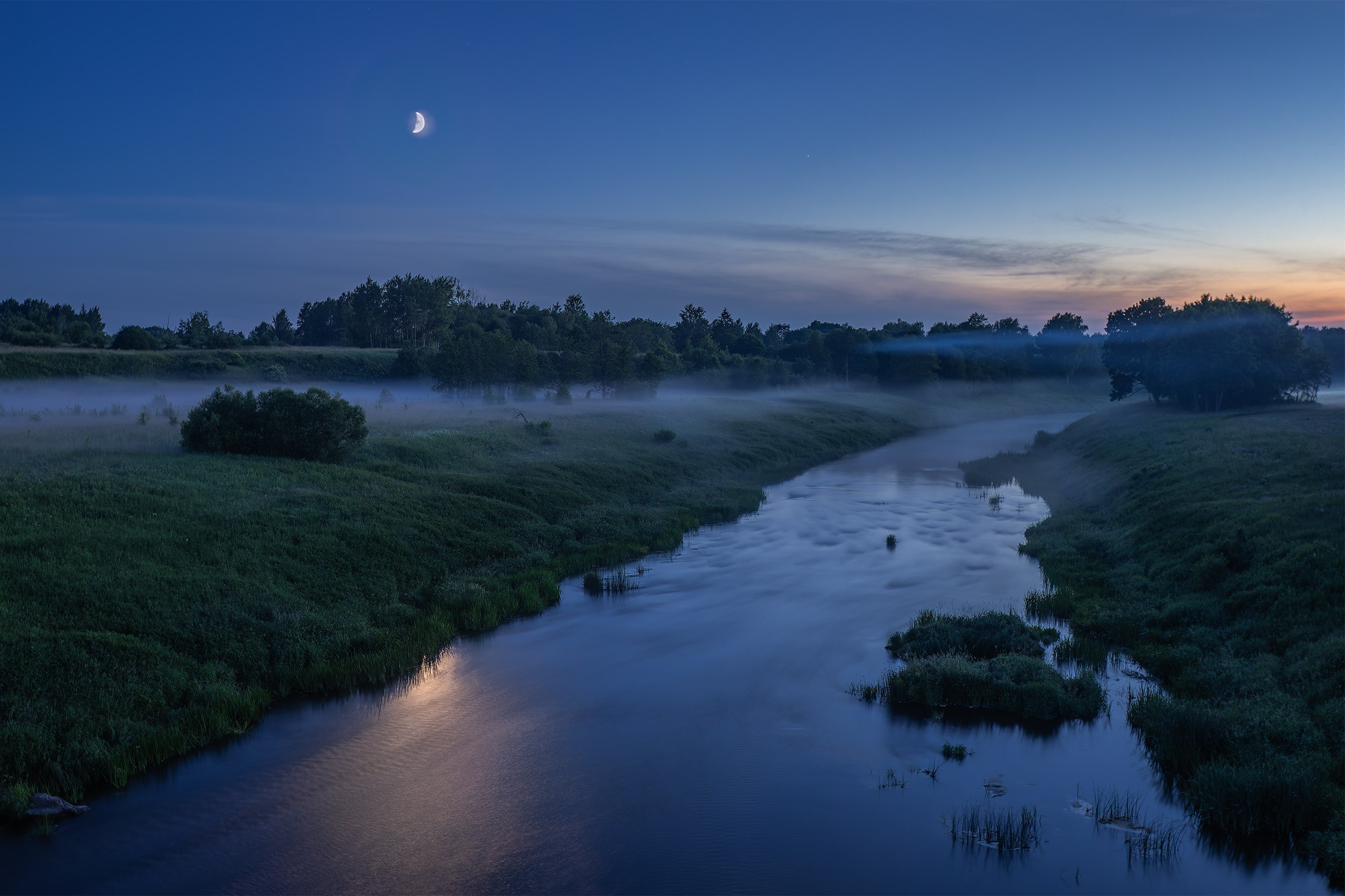 A pale moon above the Sheshupe River with low fog hanging over the water during blue hour in East Prussia.