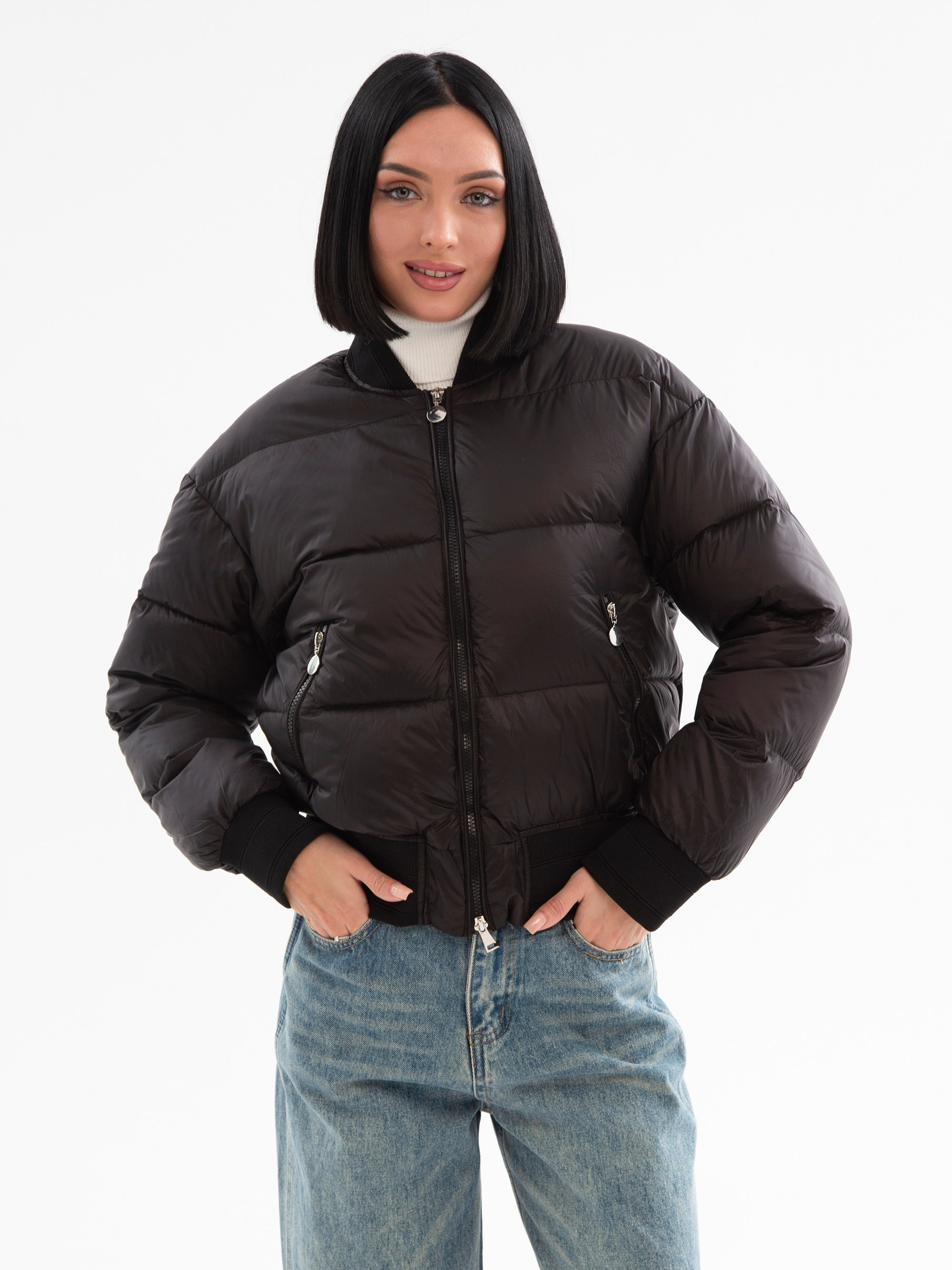 Девушка брюнетка в черной куртке и джинсах стоит на белом фоне