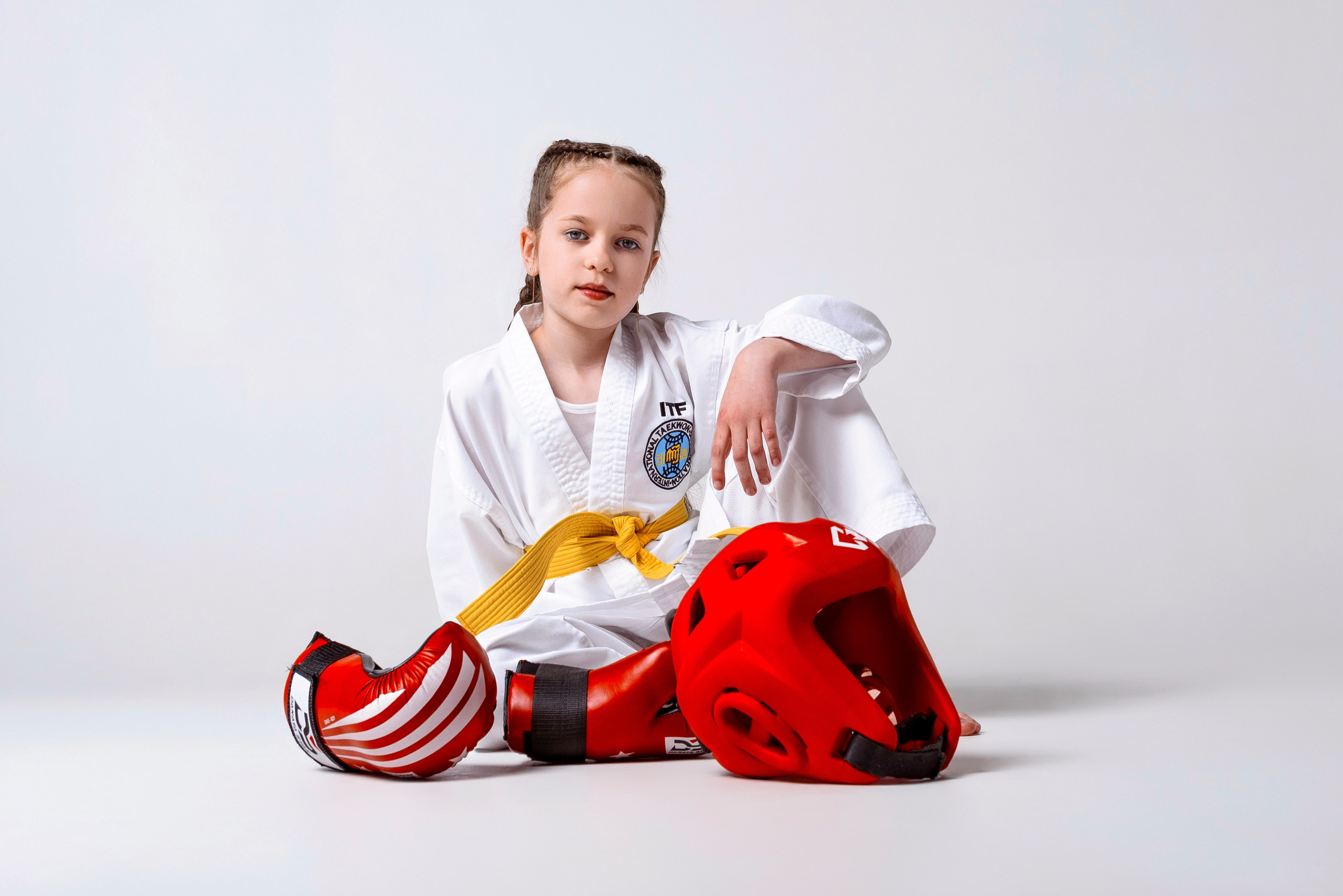 На изображении изображено лицо, одетое в белую форму Тхэквондо (добок) с желтым поясом. Человек носит красные боксерские перчатки и, вероятно, готовится к тренировке боевых искусств. На форме есть нашивки, одна из которых содержит буквы "ITF", что, вероятно, указывает на Международную федерацию Тхэквондо. Фон светлый и однородный, что обеспечивает четкий обзор объекта.