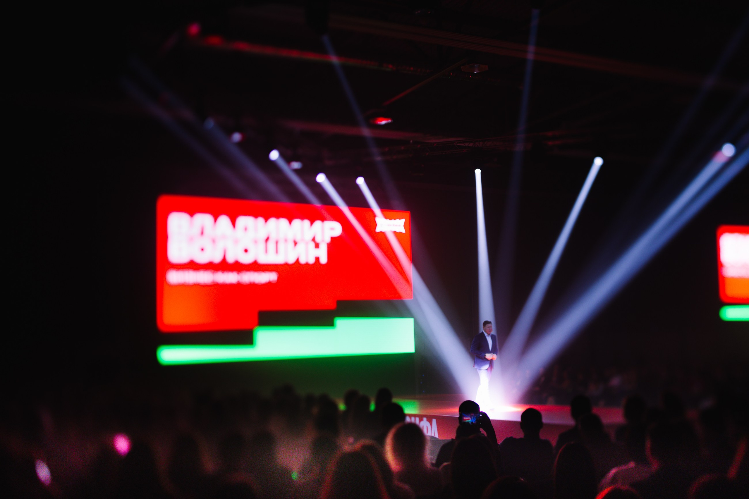 Экран и лучи: Яркие световые лучи и большой экран с логотипом «Альфа Конфа» в затемненном зале.