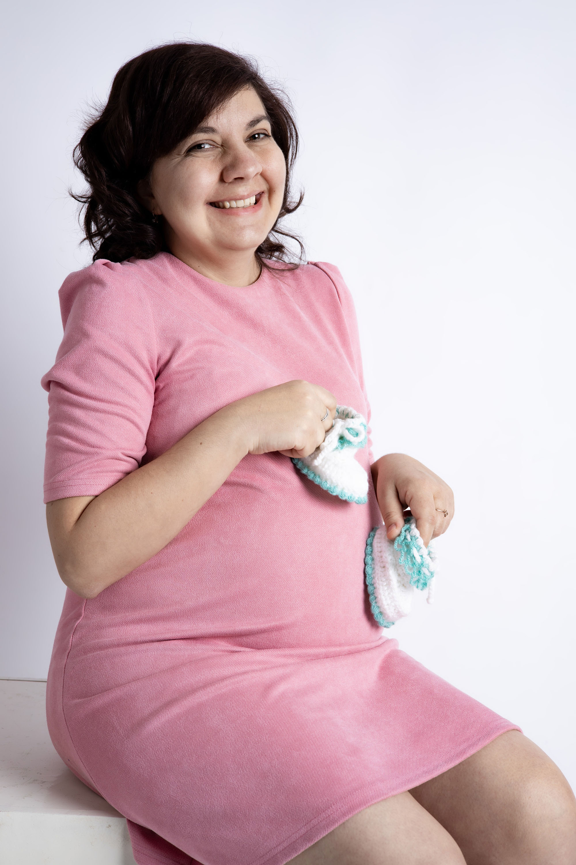 Беременная женщина сидит, в руках придерживает пинетки на животе. Фотосессия для беременных  Астрахань.