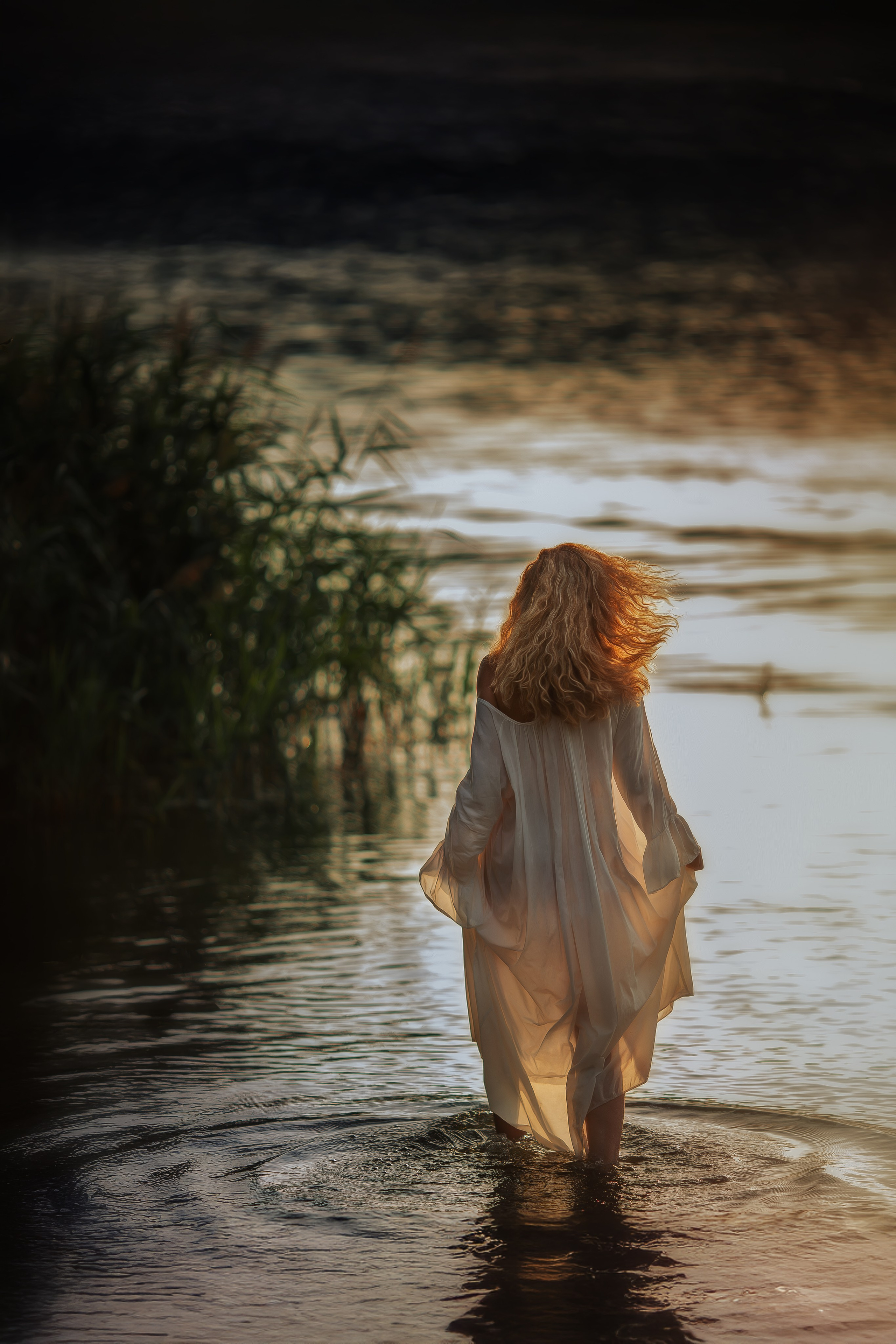 Женщина с длинными вьющимися волосами в светлом прозрачном платье идёт по воде навстречу закатному свету, ветер развивает её волосы, вода вокруг играет холодными и тёплыми оттенками.
