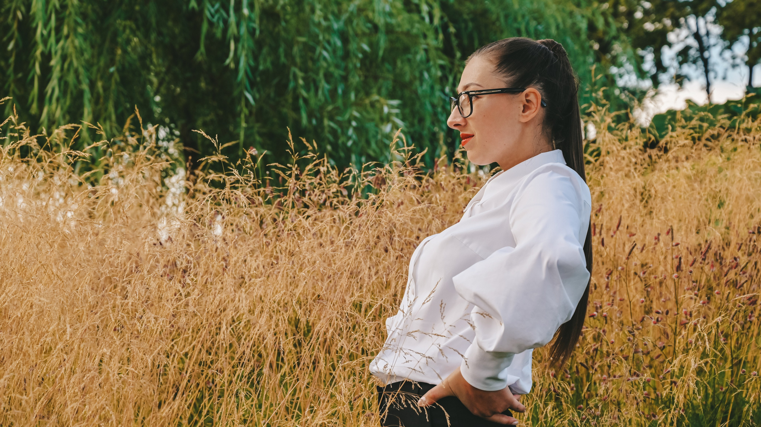 Девушка в деловом и элегантном образе с белой блузкой, черными очками и наушниками стоит на фоне живописного зеленого ландшафта. Ее длинные волосы аккуратно уложены в высокий хвост, что придает ей современный и профессиональный вид. Она одета в строгий комплект, который идеально сочетается с окружающей природной средой. Светлый фон неба и яркие краски растений создают ощущение свежести и легкости. Этот снимок демонстрирует мастерство работы с освещением и композицией, делая акцент на игре света и тени, а также на гармоничном сочетании человека и природы. Perfect for business and lifestyle photography enthusiasts!