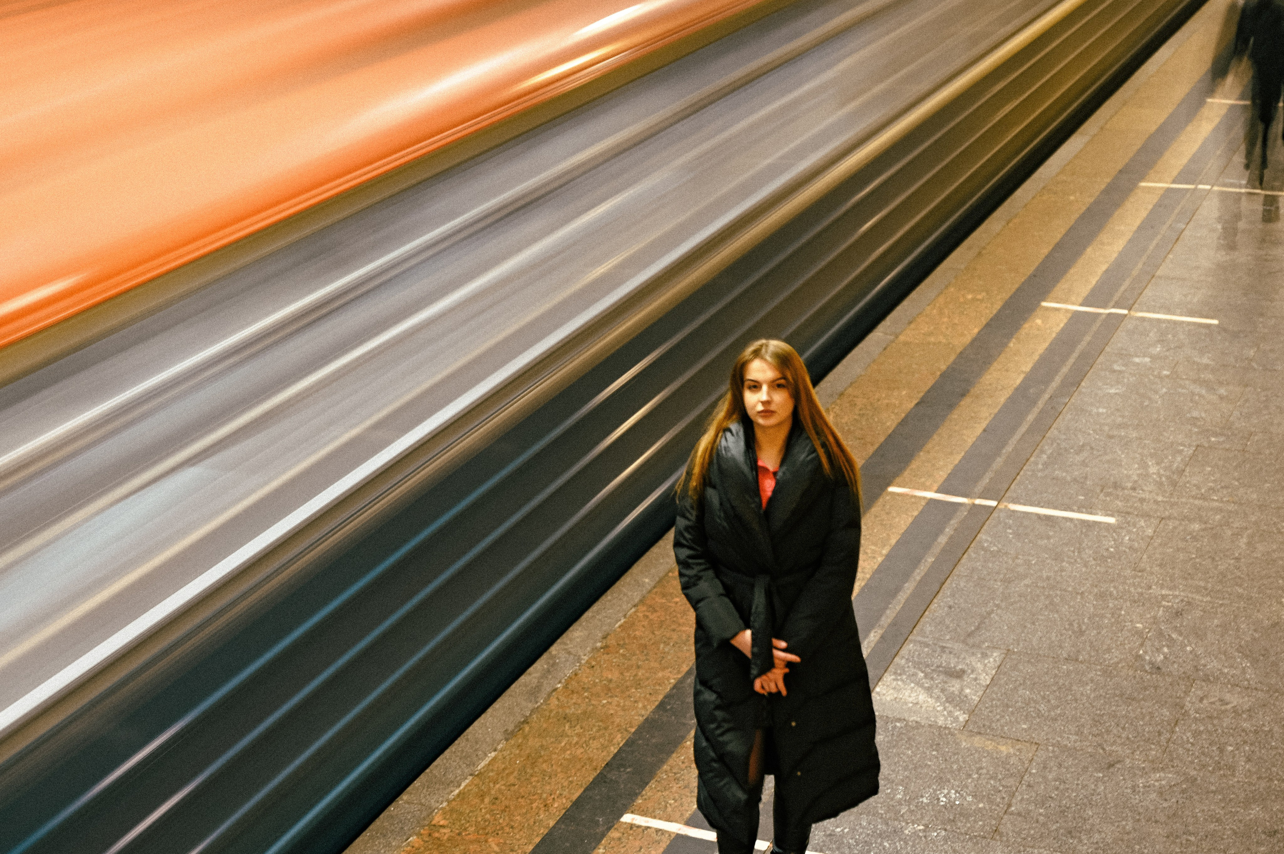Молодая женщина в темном пуховике стоит на платформе станции метро. Она имеет длинные волосы и смотрит прямо в камеру, демонстрируя уверенность и сосредоточенность. Поезд, движущийся мимо, создает эффект размытия горизонтальных линий, что подчеркивает динамику городской среды. Освещение станции яркое, акцентирующее внимание на модели. Женщина находится в центре кадра, а фон состоит из металлической стены платформы и полосатого рельефа от поезда. Фотография передает контраст между спокойствием модели и движением вокруг нее.