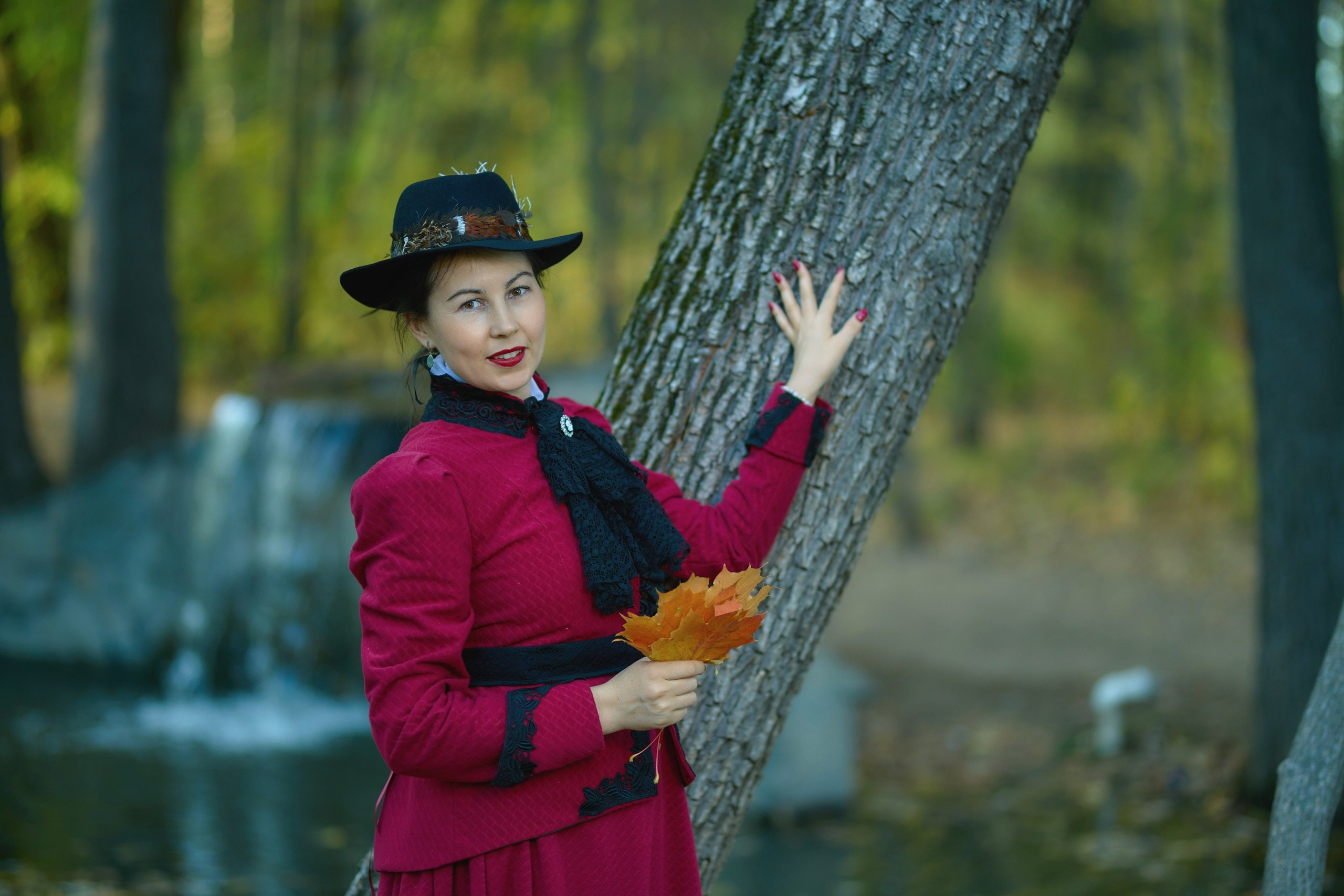 Женщина в шляпе, красном платье и жакете стоит на берегу пруда