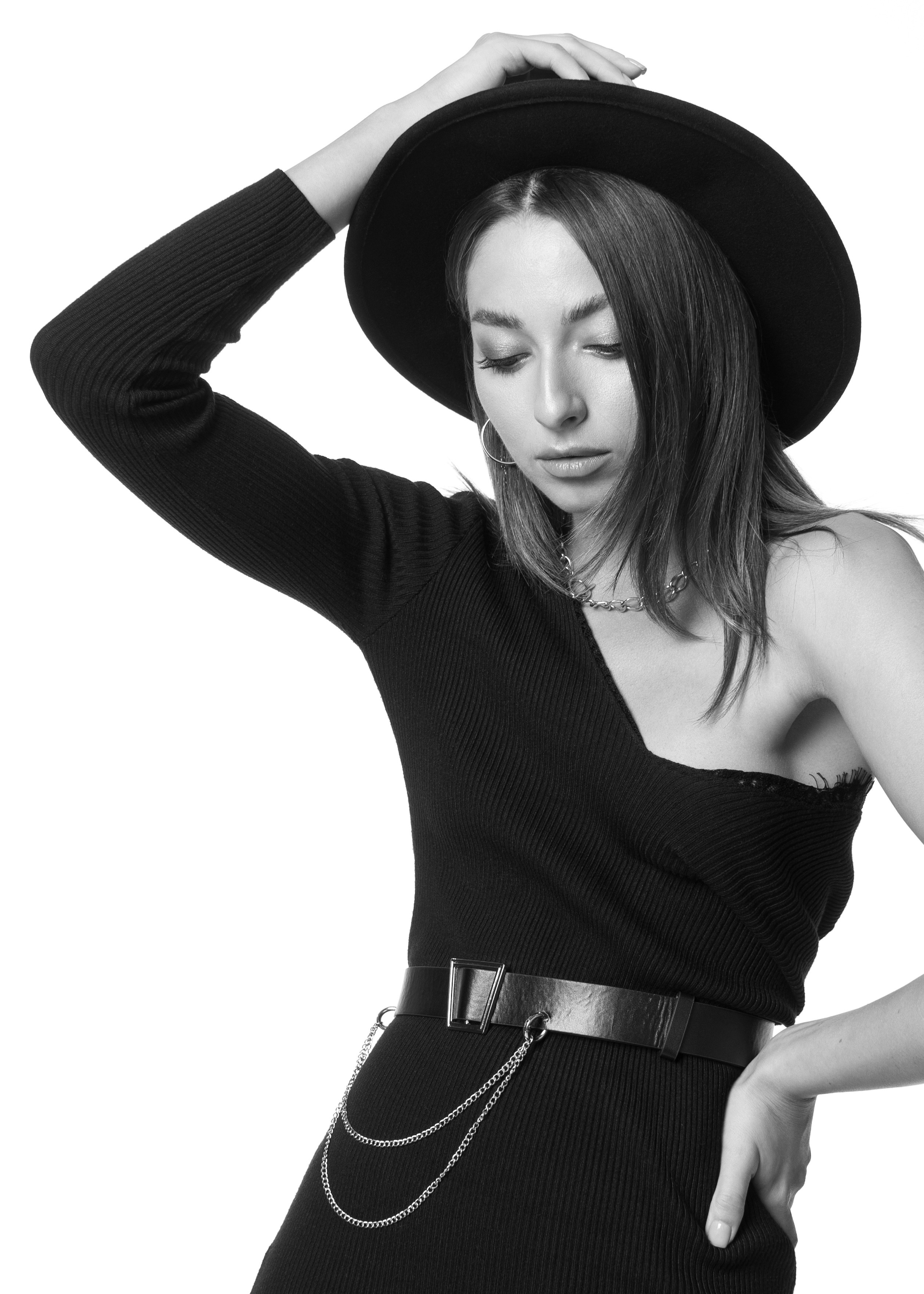 Красивая молодая девушка с тёмными волосами в черном платье с оголённым плечом и цепочкой на талии в фетровой шляпе чёрно-белое фото