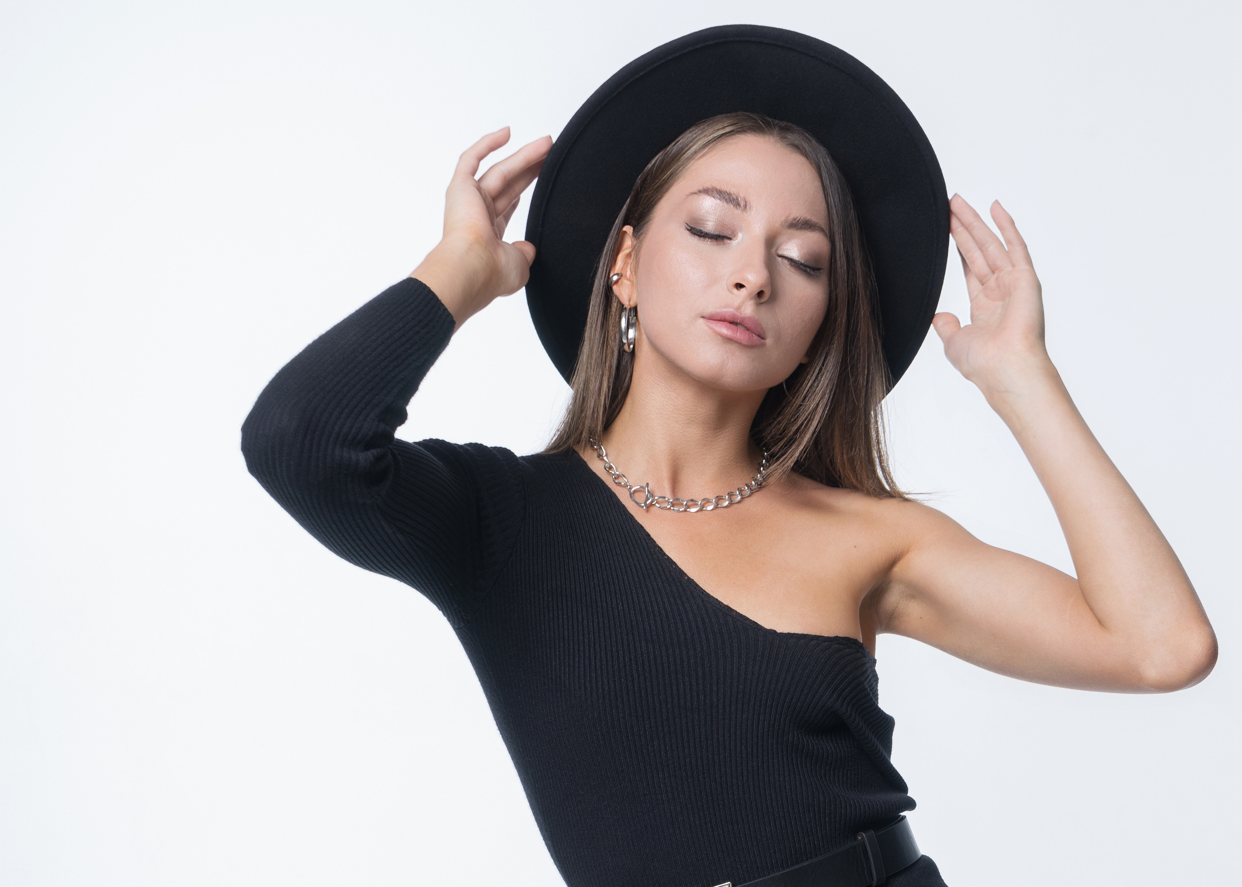 Красивая молодая девушка с тёмными волосами в черном платье с оголённым плечом и цепочкой на талии в фетровой шляпе кадрирование