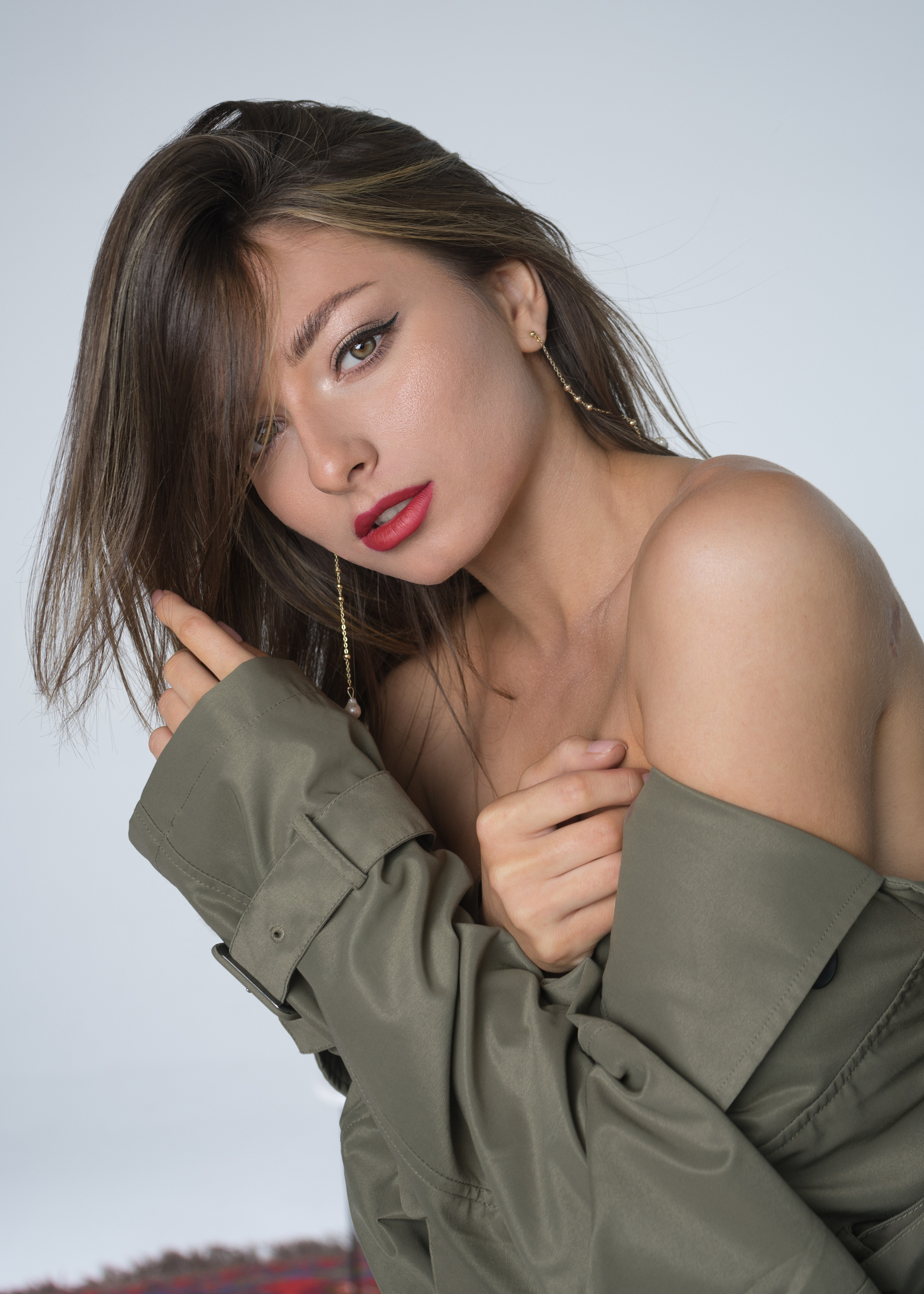 Красивая молодая девушка с тёмными волосами с красной губной помадой на губах в тренче цвета хаки с оголённым плечом