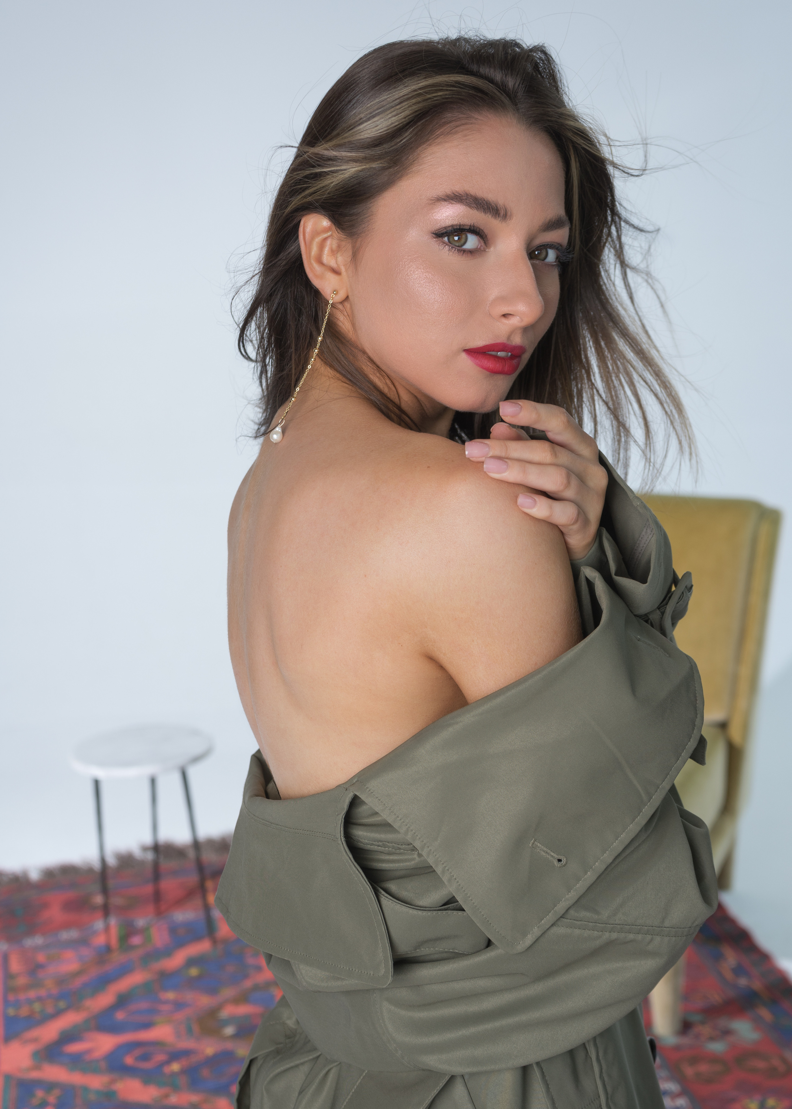 Красивая молодая девушка с тёмными волосами с красной губной помадой на губах в тренче цвета хаки с оголённым плечом и открытой спиной
