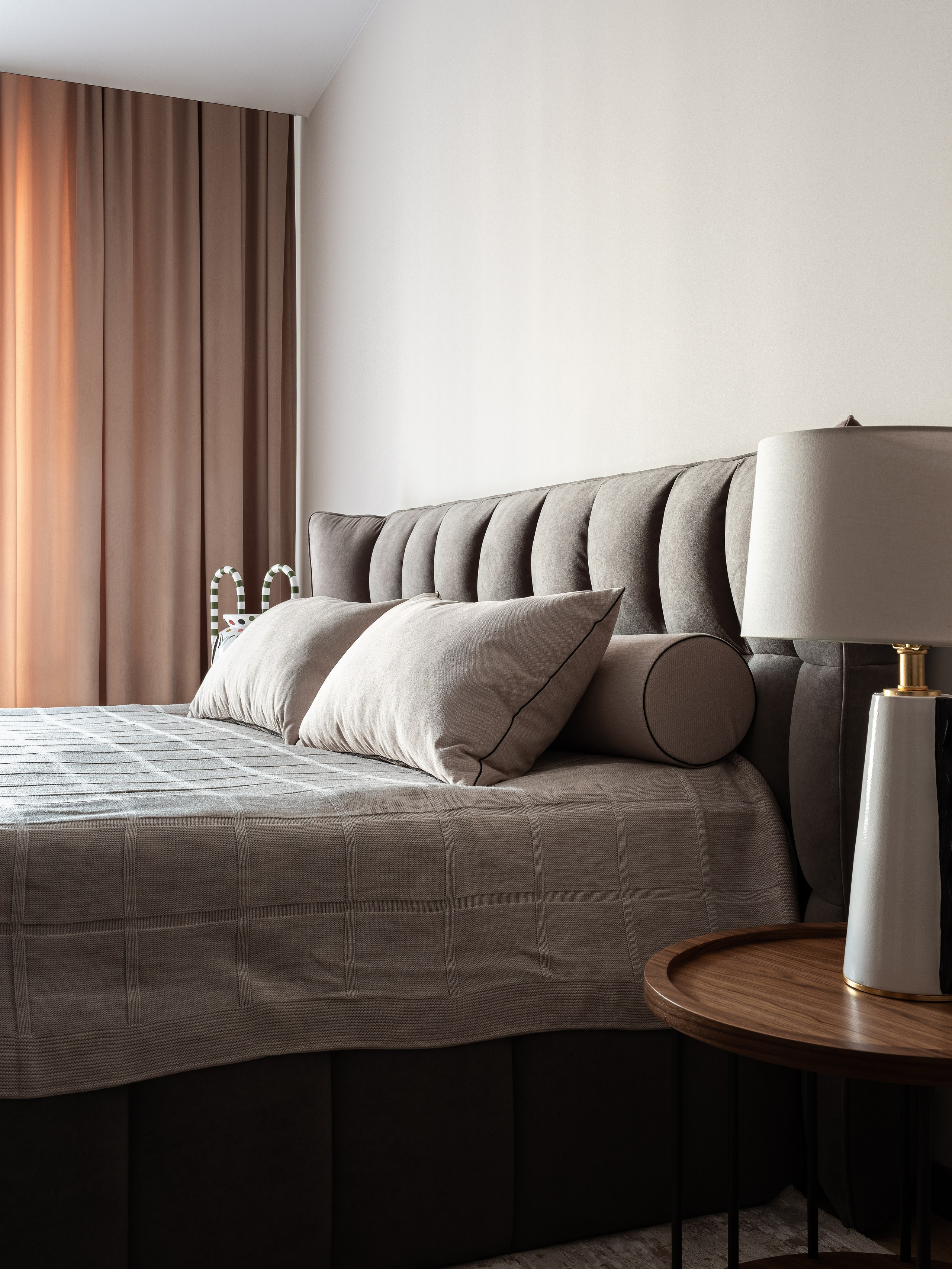 Уютная современная спальня с серым текстильным изголовьем, прикроватным столиком из дерева и светильником в минималистичном стиле, дизайнер Екатерина Мищенко, фотограф Сергей Денисенко, гармония теплых штор и спокойных оттенков.