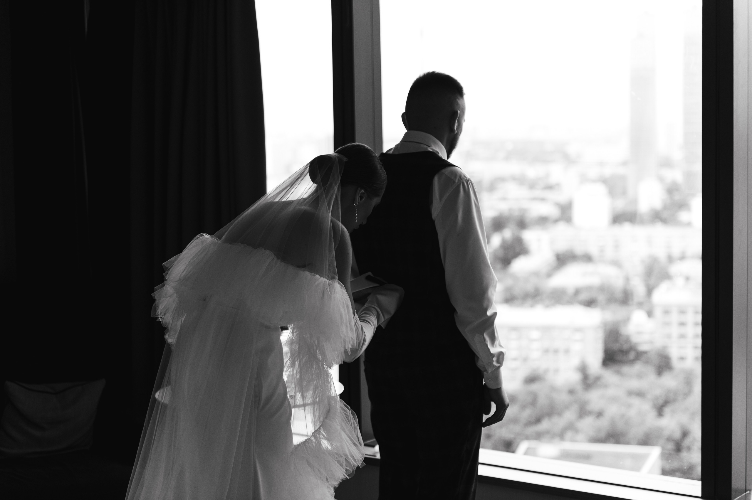 Невеста поправляет костюм жениха около окна в отеле.