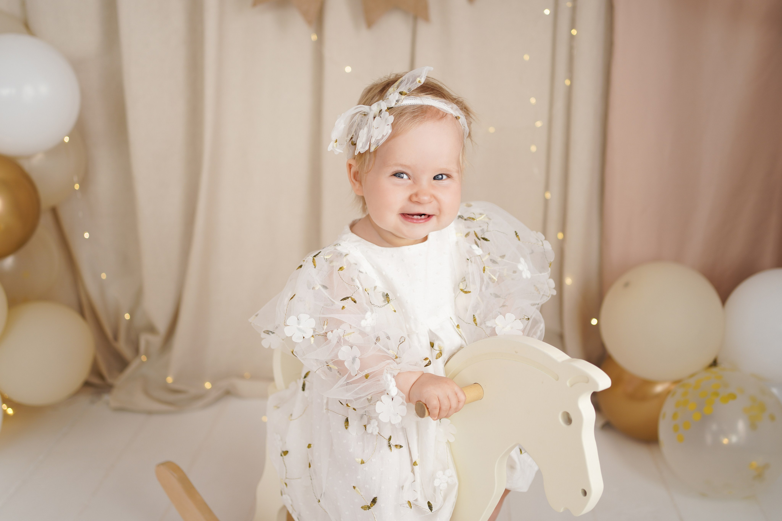 Малышка в белом платье  катается на игрушечной лошадке.