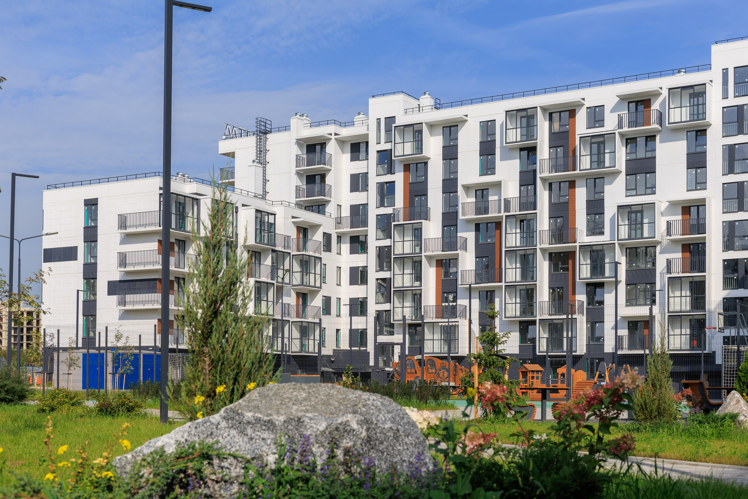 На изображении представлена застройка современного жилого комплекса. В центре композиции находятся два жилых многоквартирных дома, окруженные небольшим количеством зелени и цветов. Стиль зданий современный, с преобладанием белого цвета в отделке фасадов.
