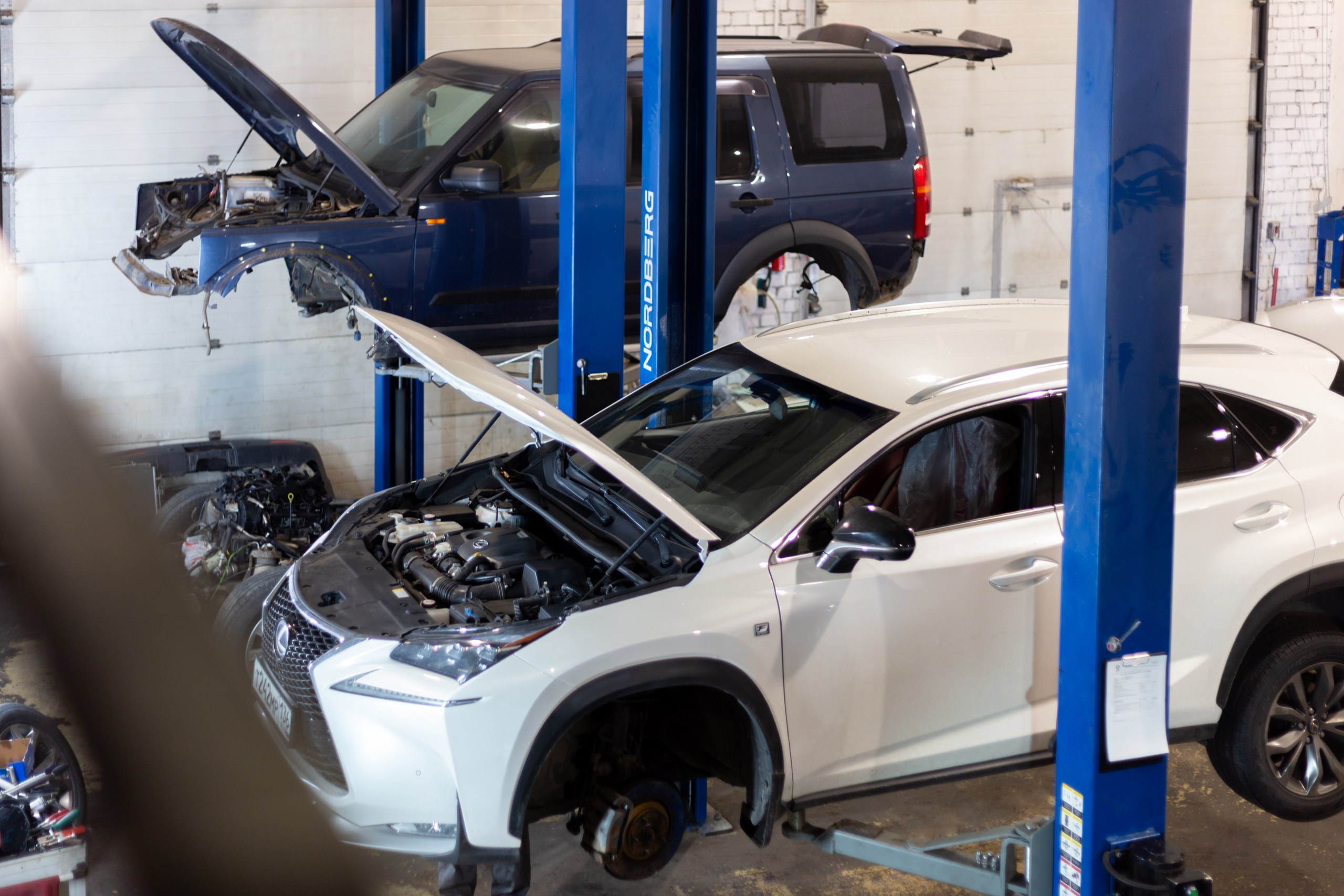 Вид сверху на белый и черный автомобиль на подъемниках в автосервисе. Белая машина с открытым капотом.