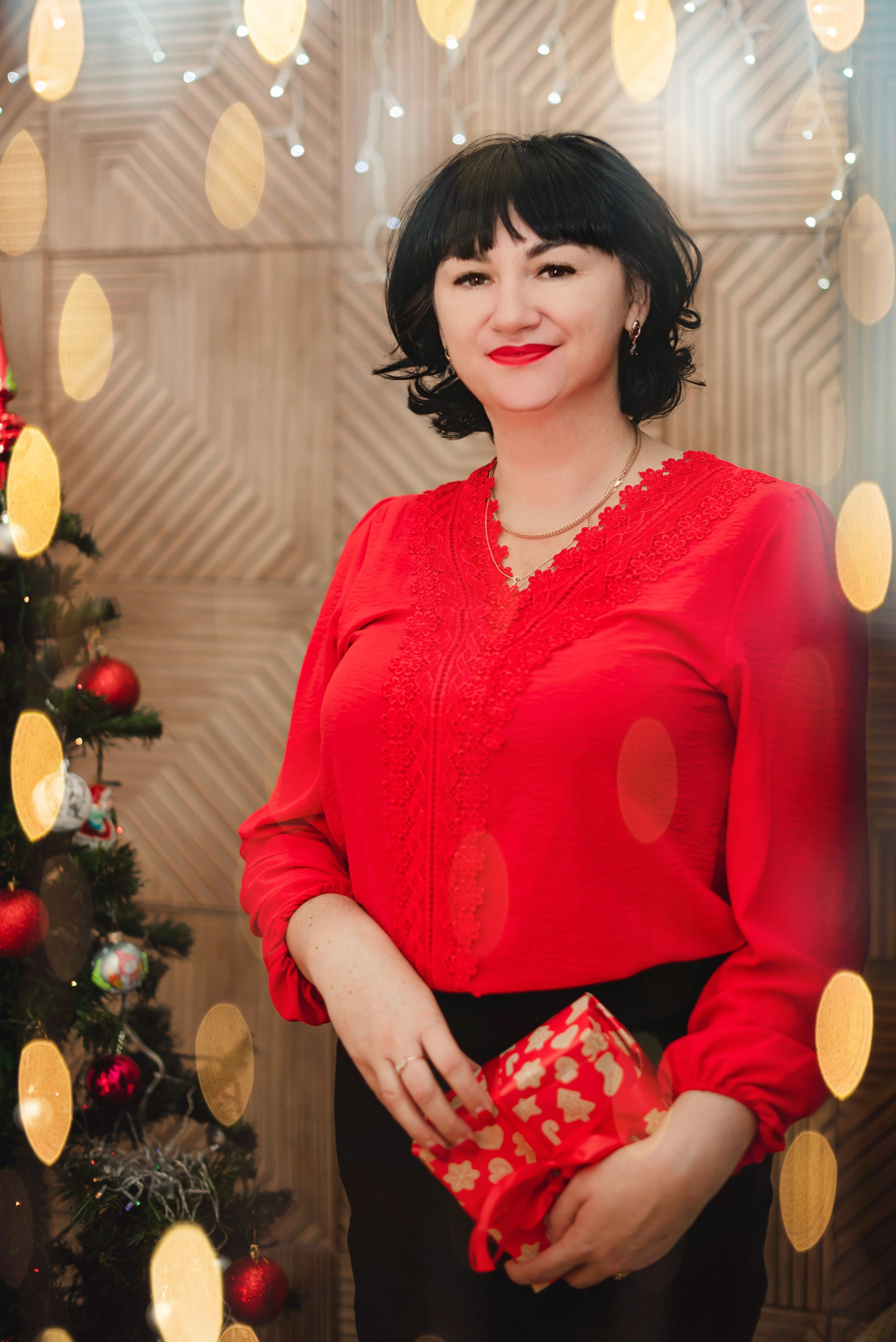 На фото женщина с подарком на новый год в студии в красной блузке около елочки дома.