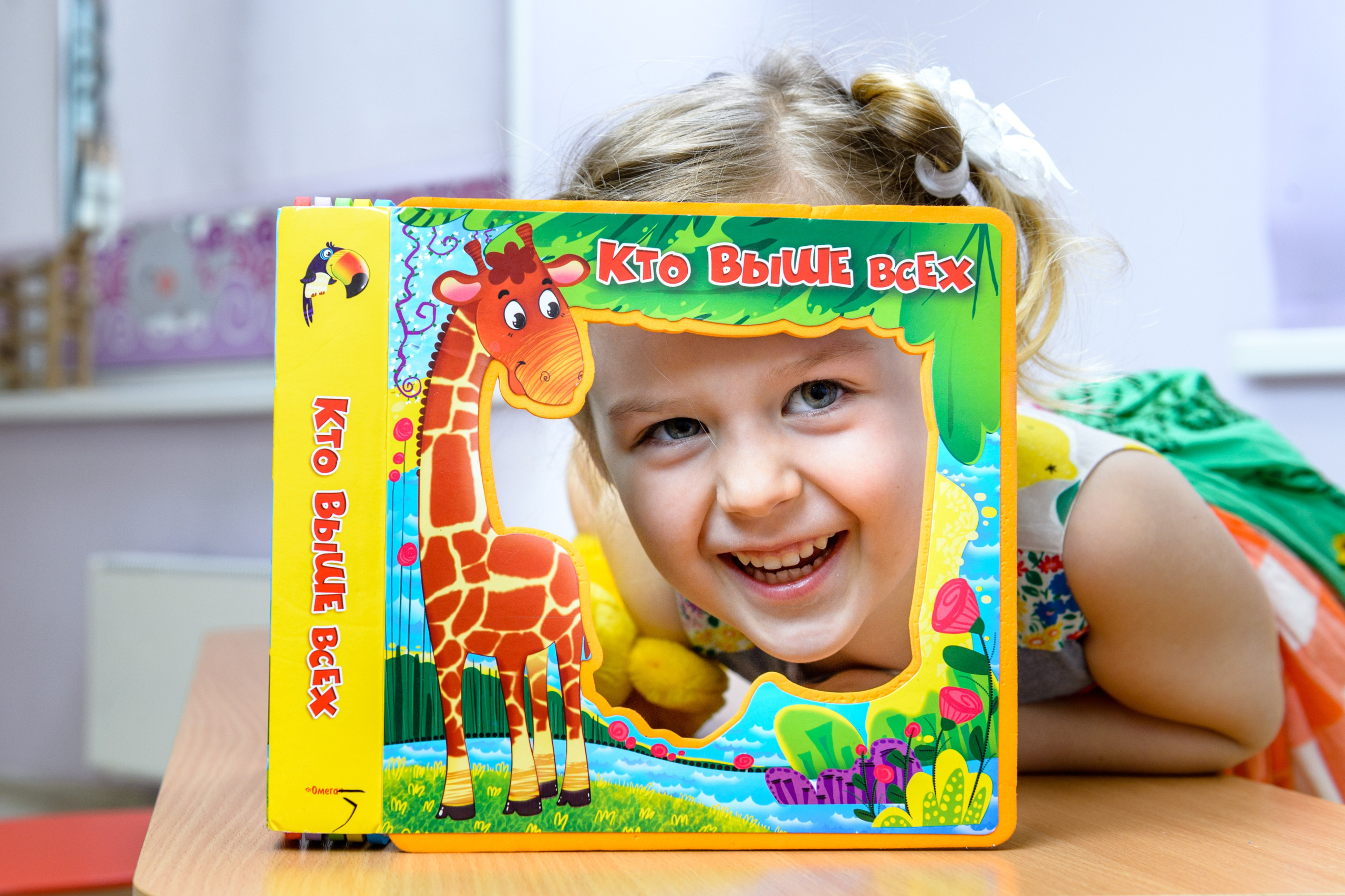 Девочка выглядывает из рамки цветной обложки детской книжки и улыбается. 