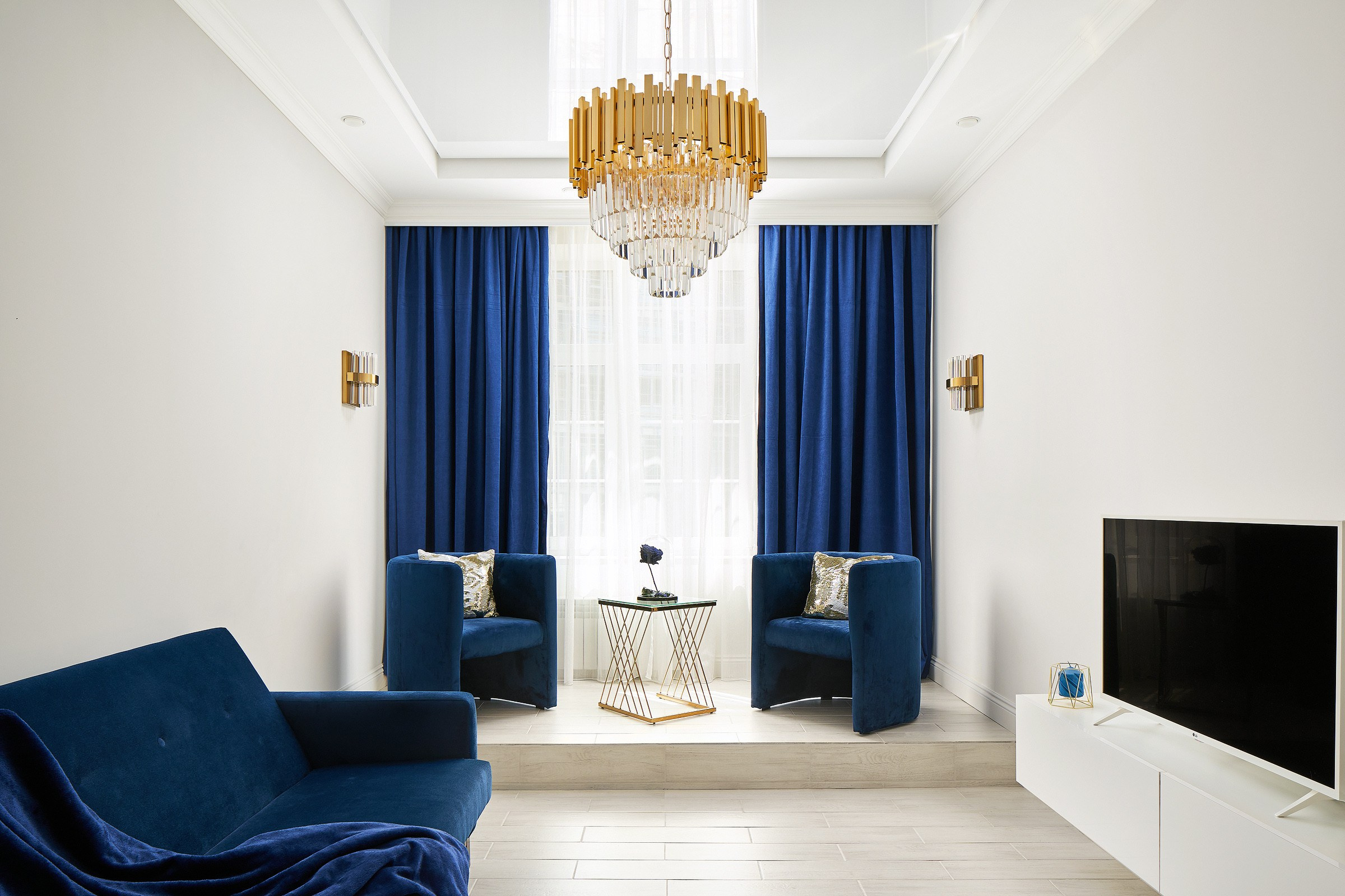 Светлая спальня-гостиная с синим диваном, синими шторами и золотистой люстрой