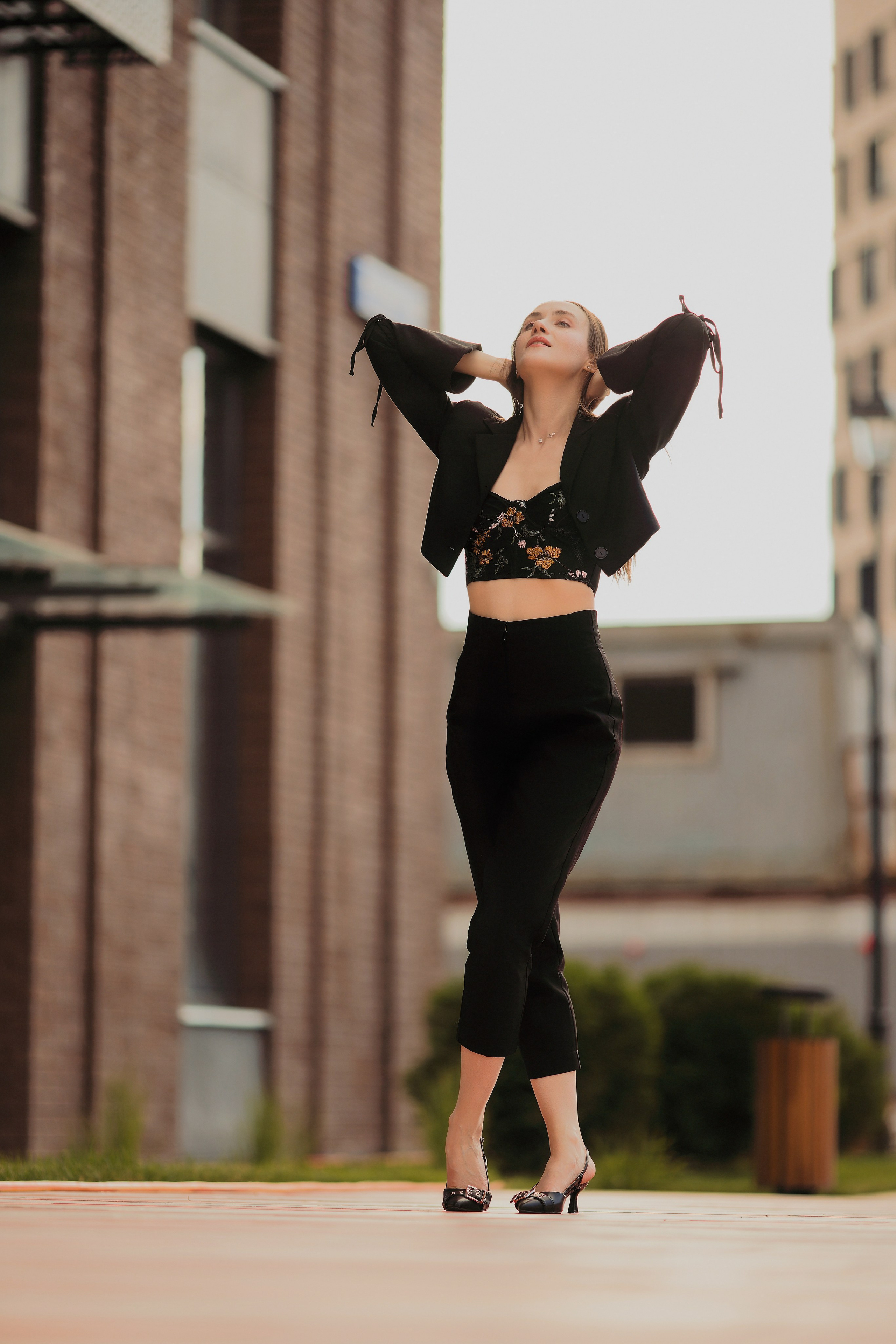 Полноростовой портрет девушки в черном костюме с пиджаком на улице, динамичная поза, городская архитектура на фоне