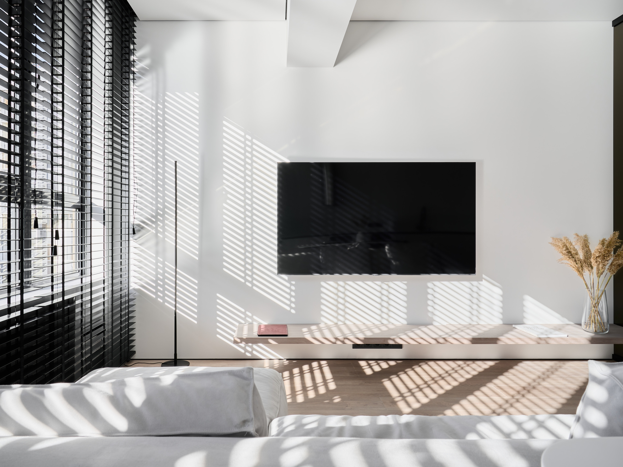 Светлая гостиная с панорамными окнами и черными жалюзи, на стене телевизор и подвесная тумба, солнечные лучи создают игру теней, дизайнер Анатолий Котов, фотограф Сергей Денисенко, стильный минимализм и уютная атмосфера.