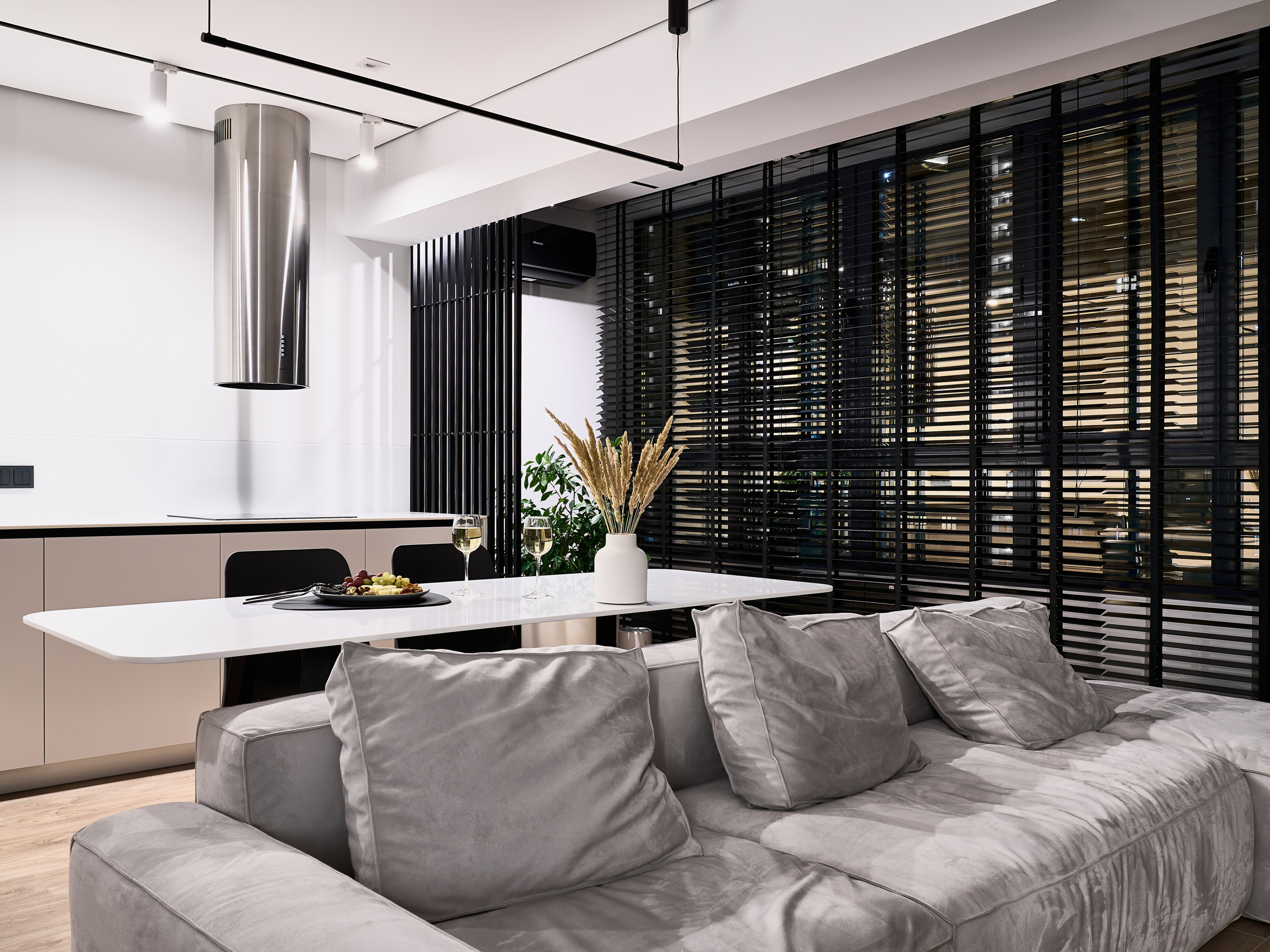 Современный интерьер с мягким серым диваном, белым столом и кухонной зоной в минималистичном стиле, выразительные черные жалюзи подчеркивают объем. Авторский дизайн Анатолия Котова, фото Сергея Денисенко.