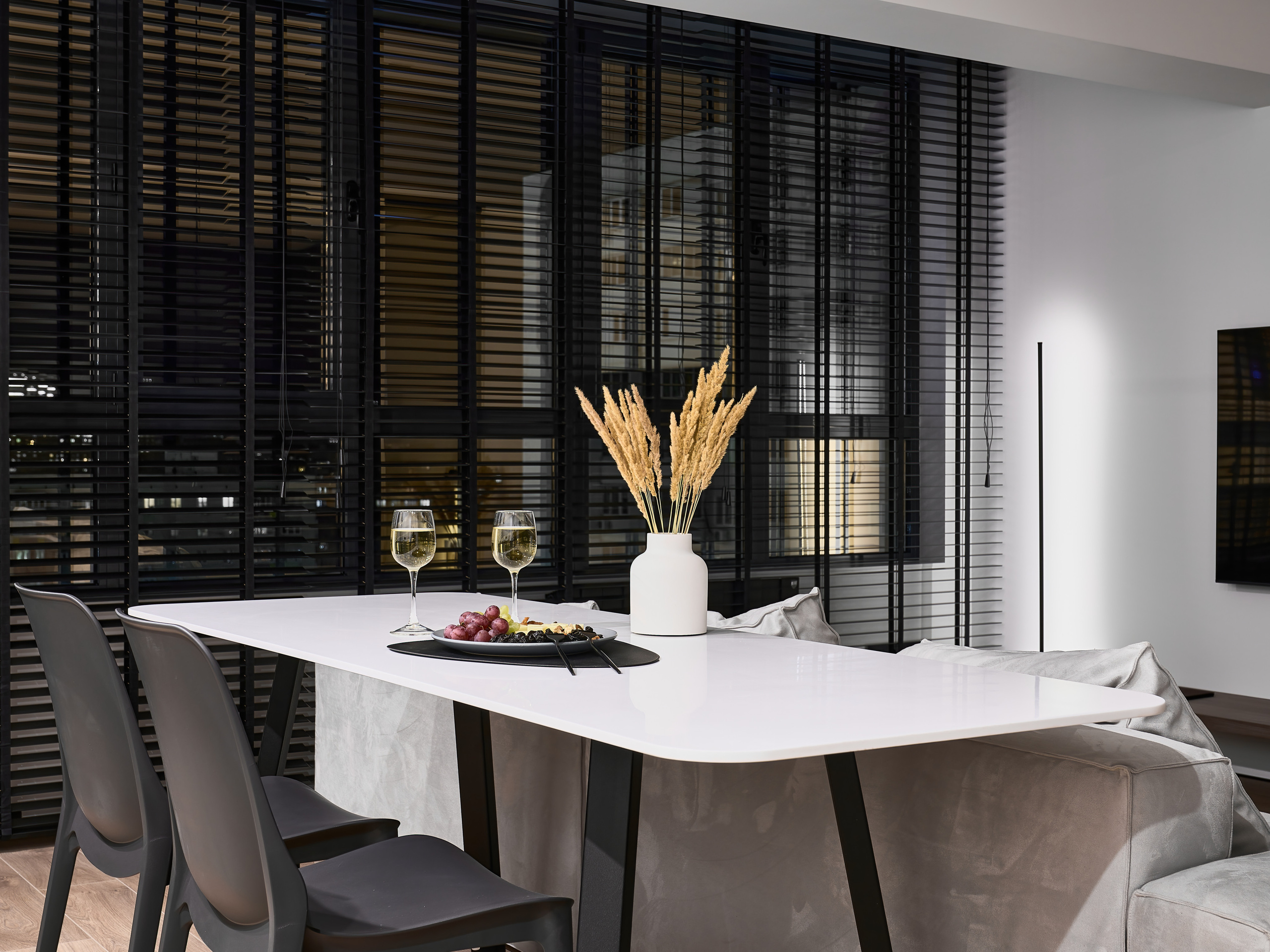 Белый обеденный стол с черными стульями, сервировка с вином и закусками, декоративная ваза с сухоцветами, на фоне панорамных окон с черными жалюзи. Дизайн Анатолия Котова, фотосъемка Сергея Денисенко.
