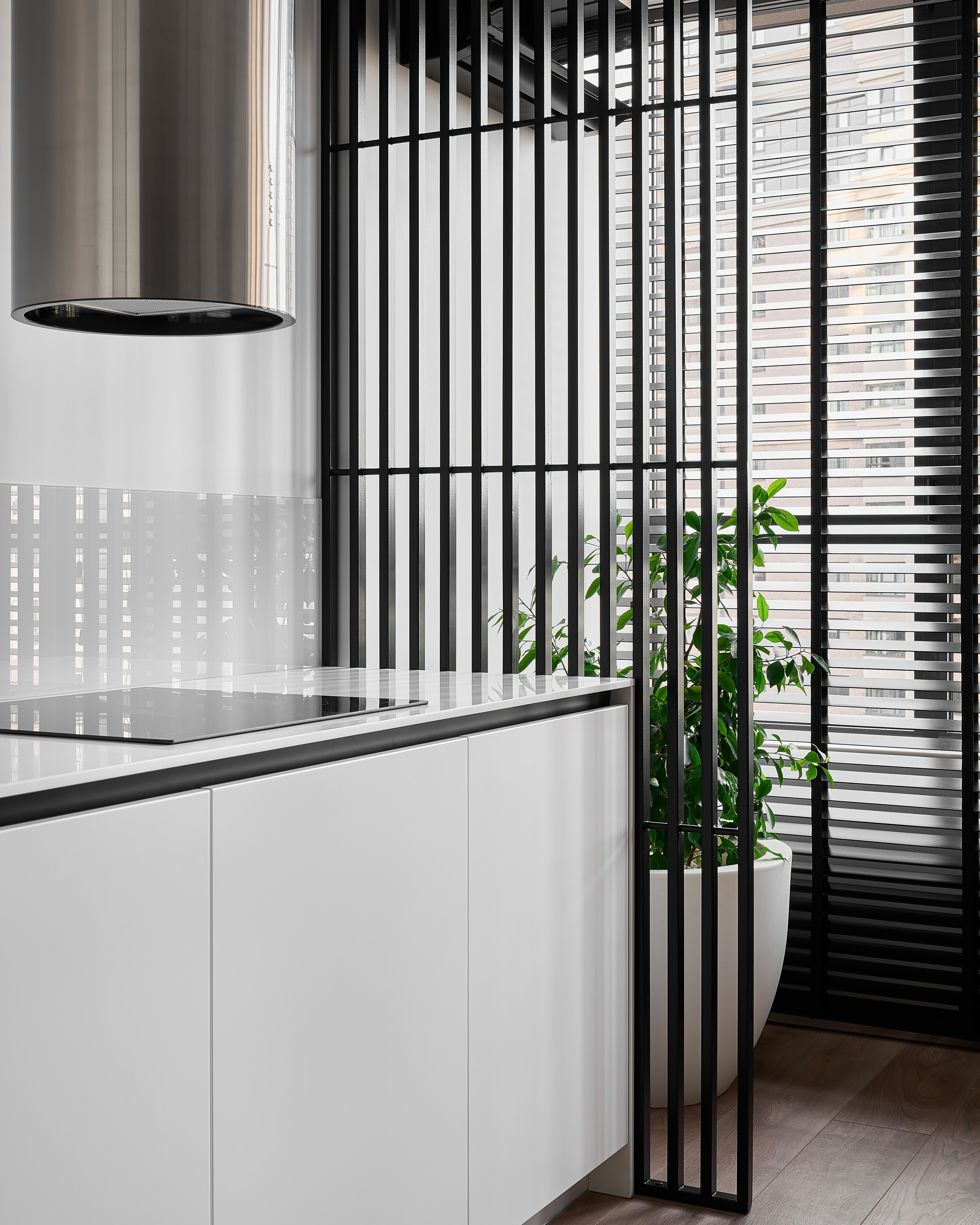 Современный интерьер кухни с белыми глянцевыми фасадами, встроенной варочной поверхностью и вытяжкой цилиндрической формы, акцент на черных вертикальных рейках и зеленом растении у окна. Дизайн Анатолий Котов, фото Сергей Денисенко.