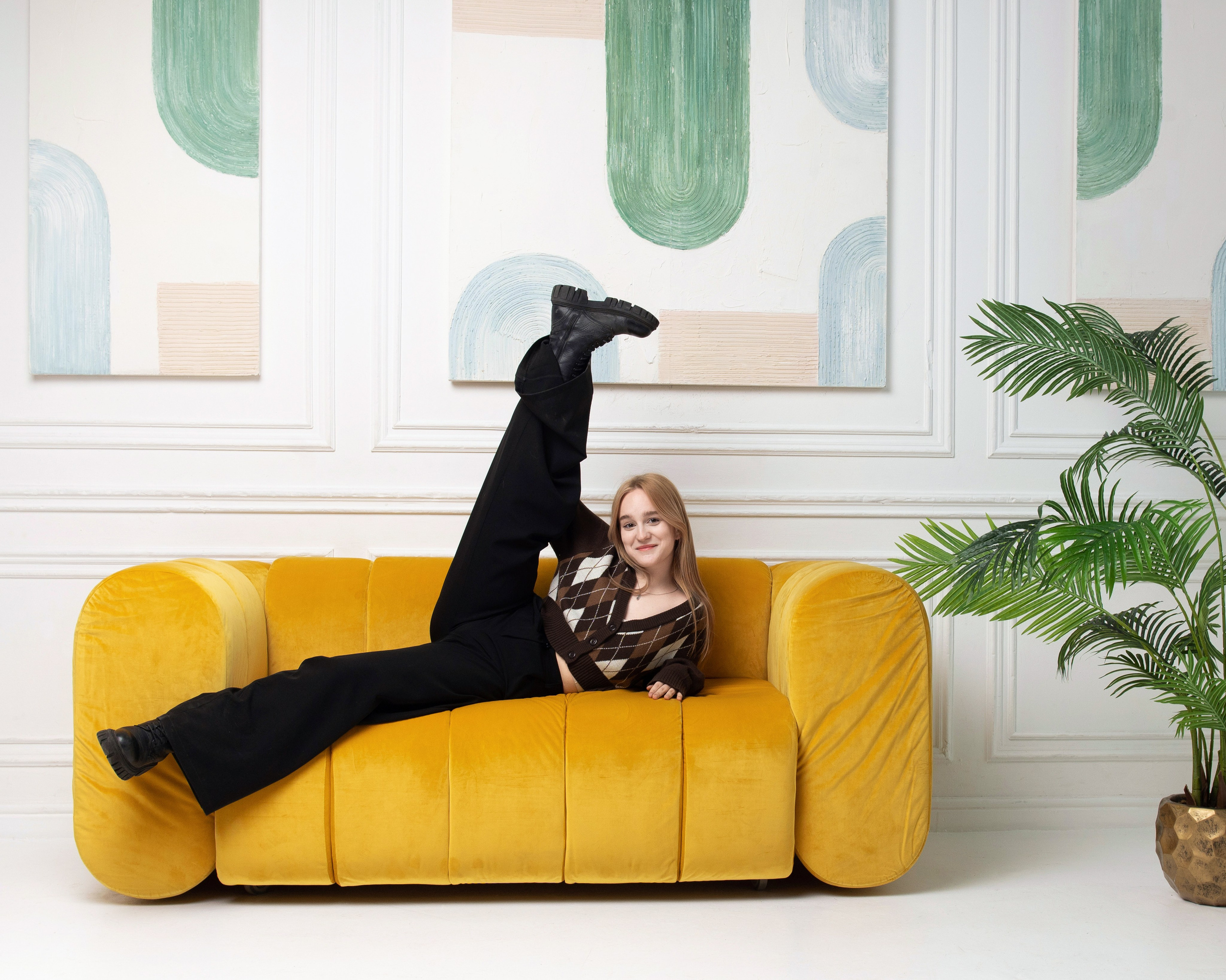 Девушка в чёрных брюках и коричневом жилете лежит на жёлтом диване в стилизованной фотостудии.