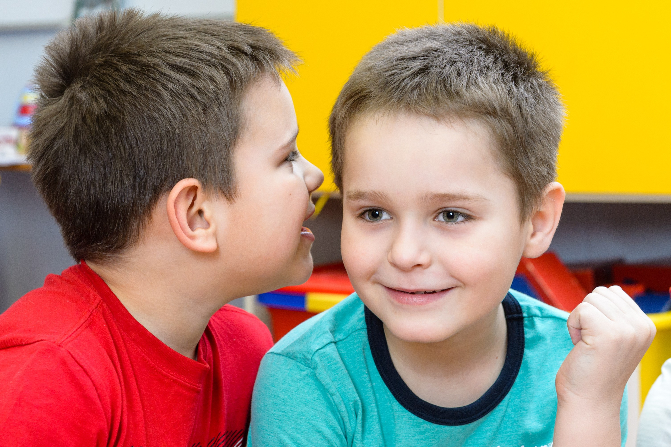 Два мальчика сидят рядом: один шепчет на ухо другому в игровом классе.