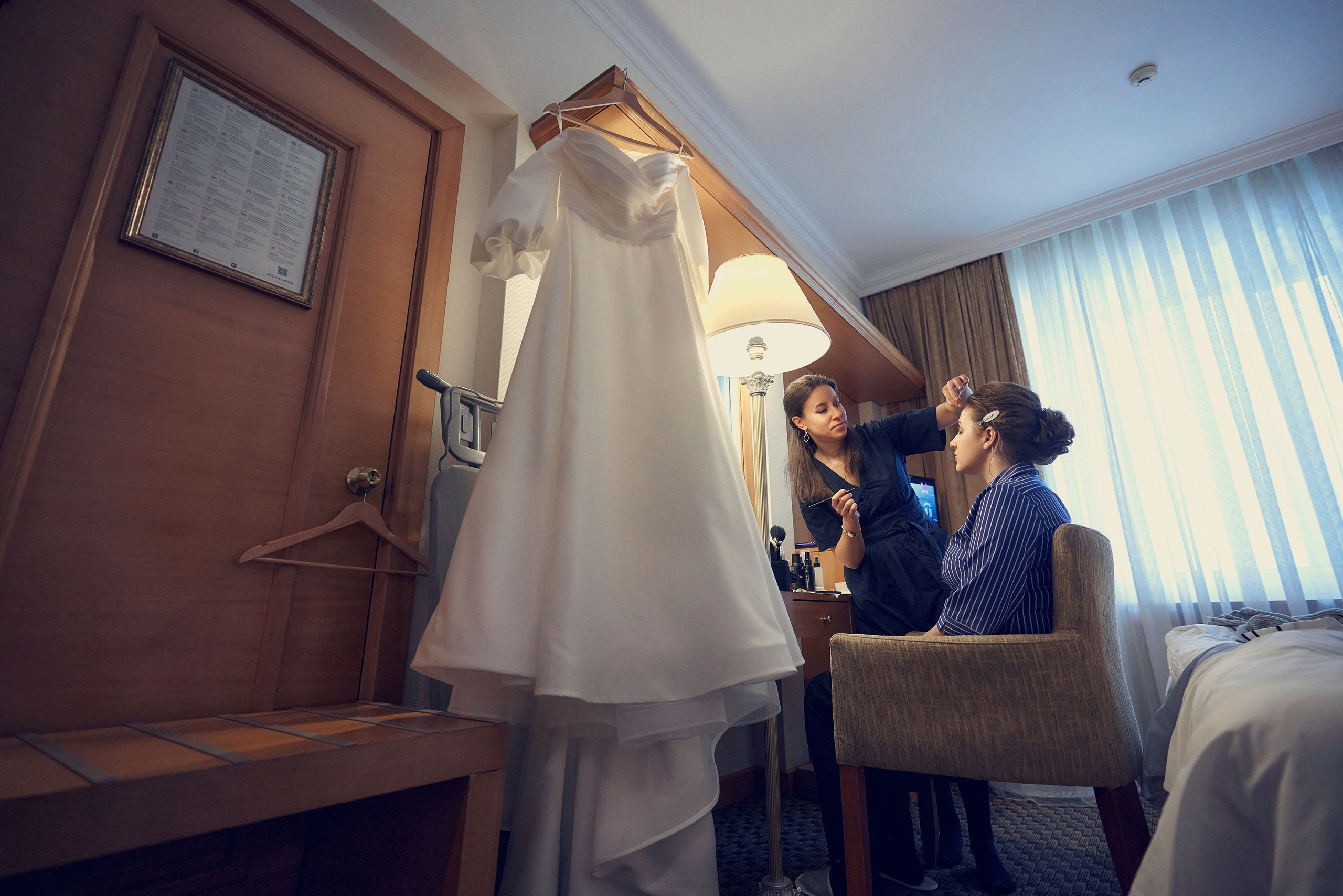 Сборы невесты в номере отеля Милан - в кадре висит платье и визажист красит невесту перед зеркалом.
