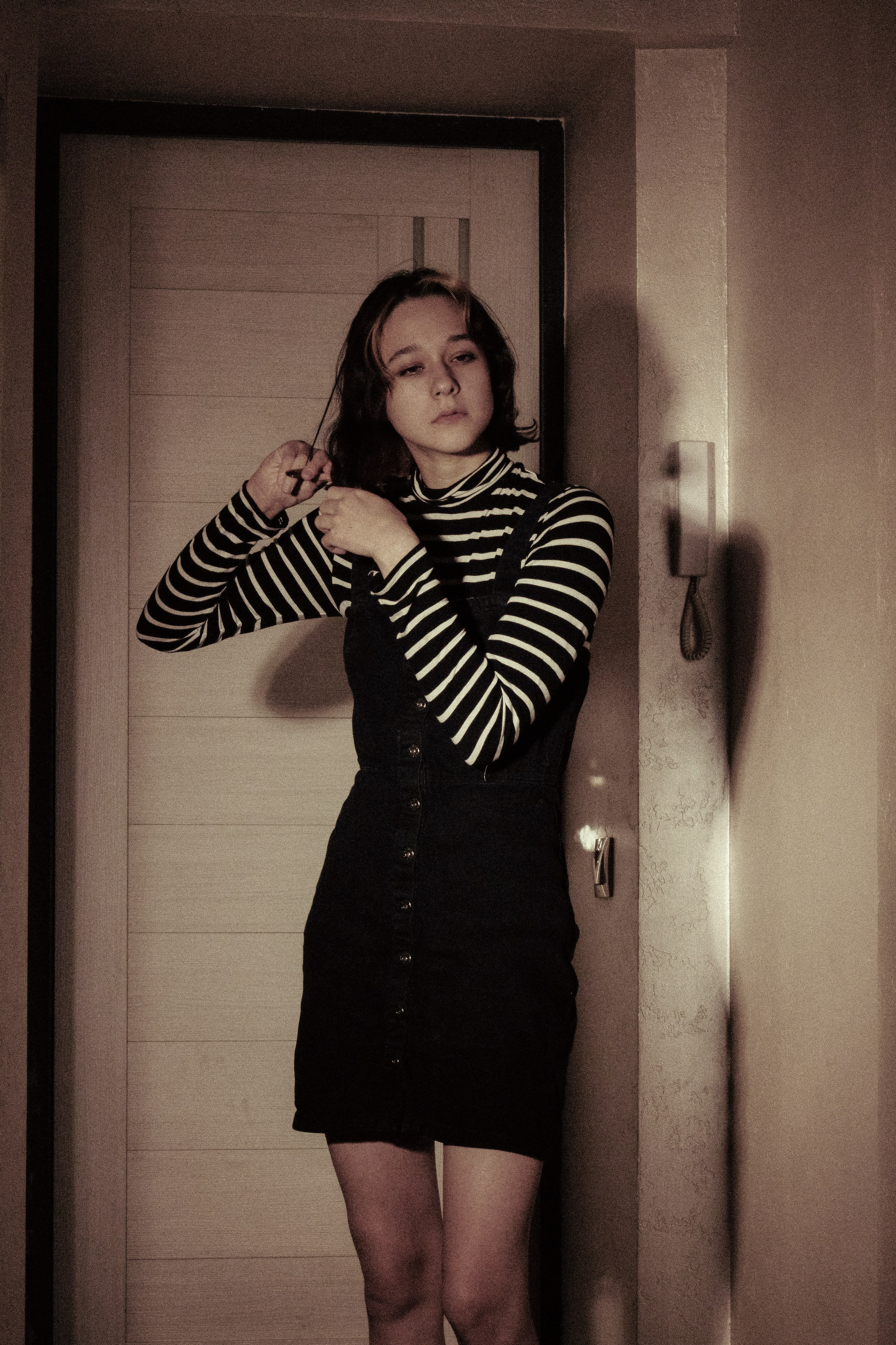 Девушка стоит перед дверью и задумчиво трогает волосы — будто не верит, что время идёт дальше.