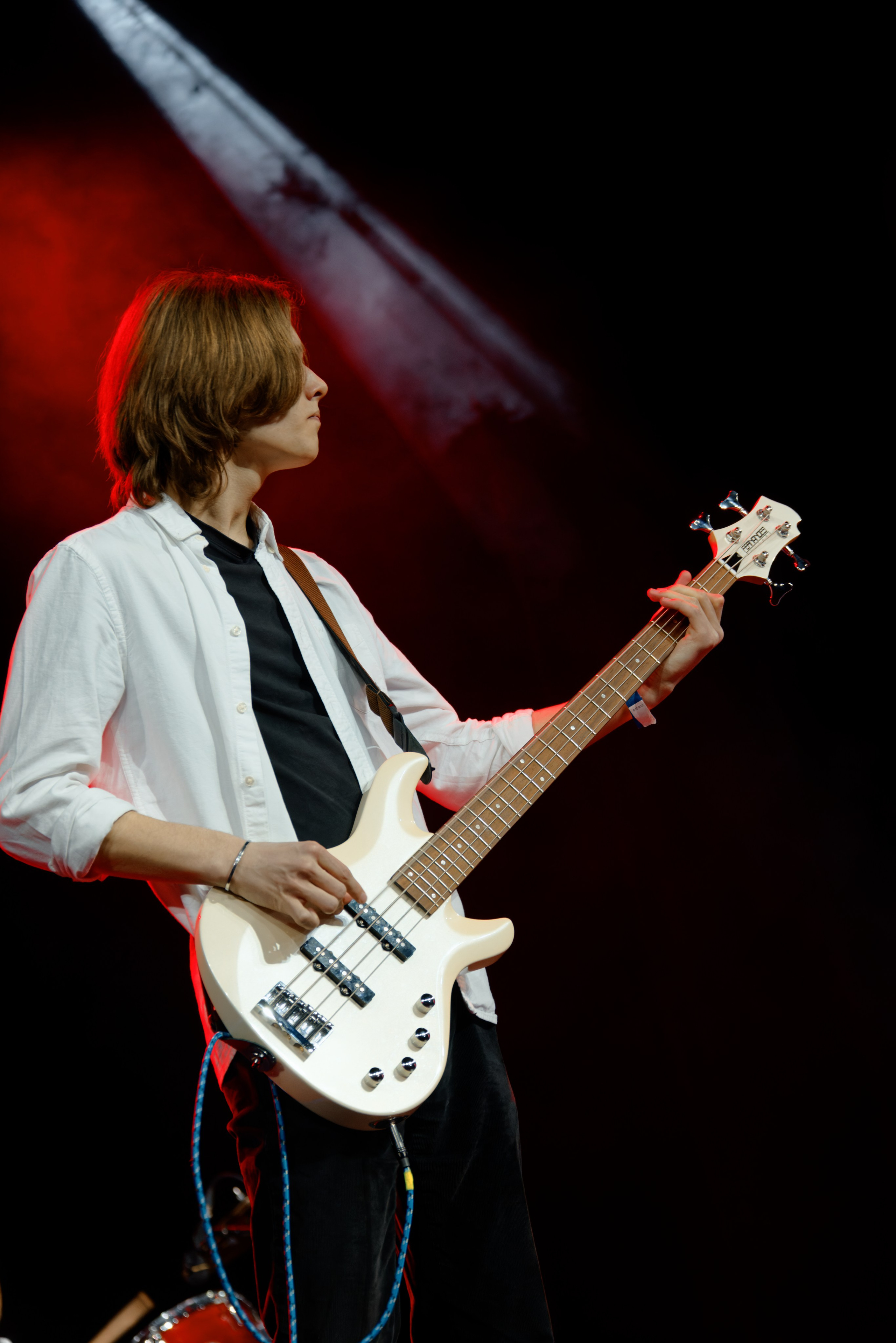 Бас-гитарист в белой рубашке играет на белой бас-гитаре на сцене с красным сценическим светом, профиль музыканта во время выступления