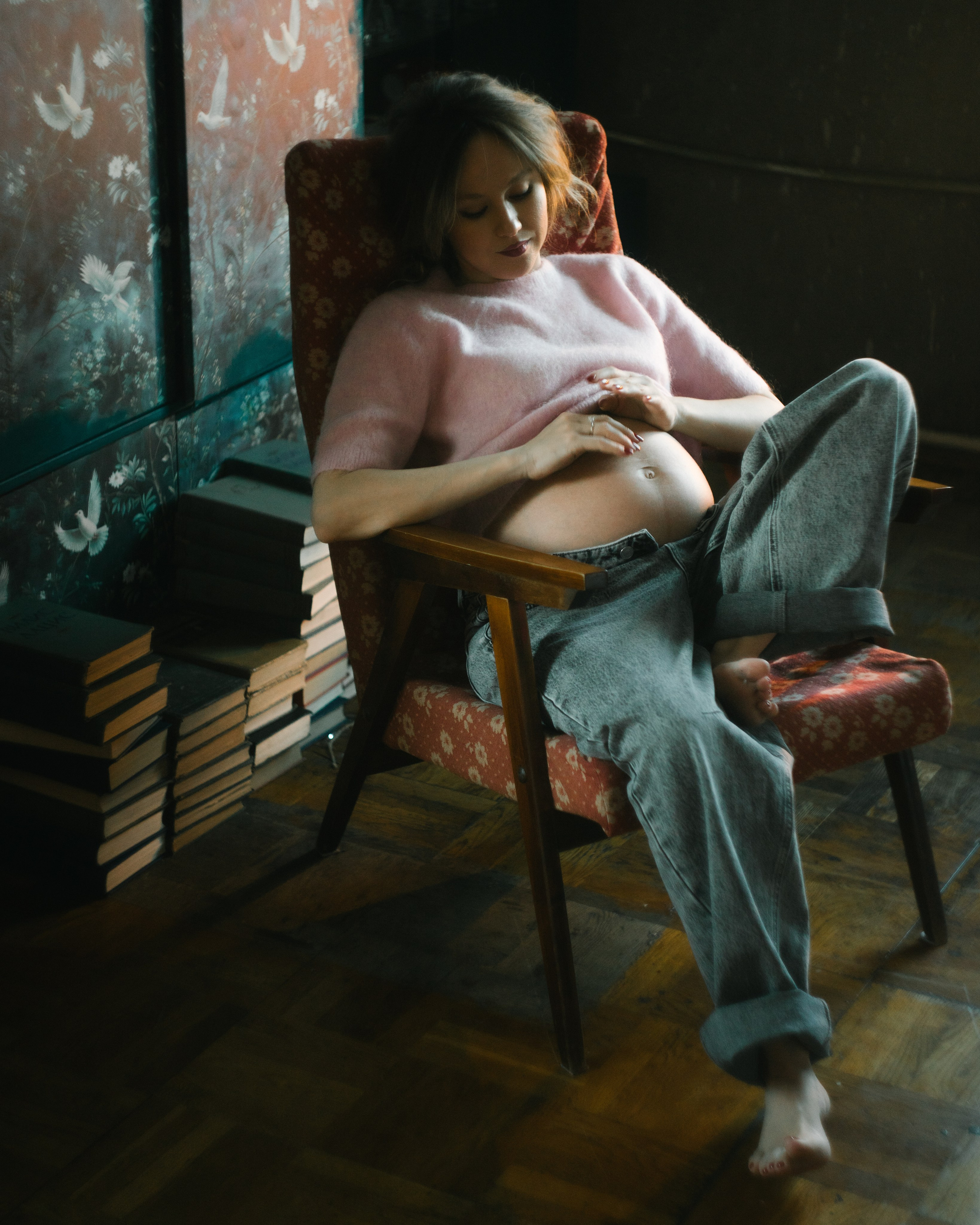 Беременная девушка в джинсах и розовом свитере в ретро-фотостудии Санкт-Петербурга: кресло, стопки книг, камерная фотосессия беременности.