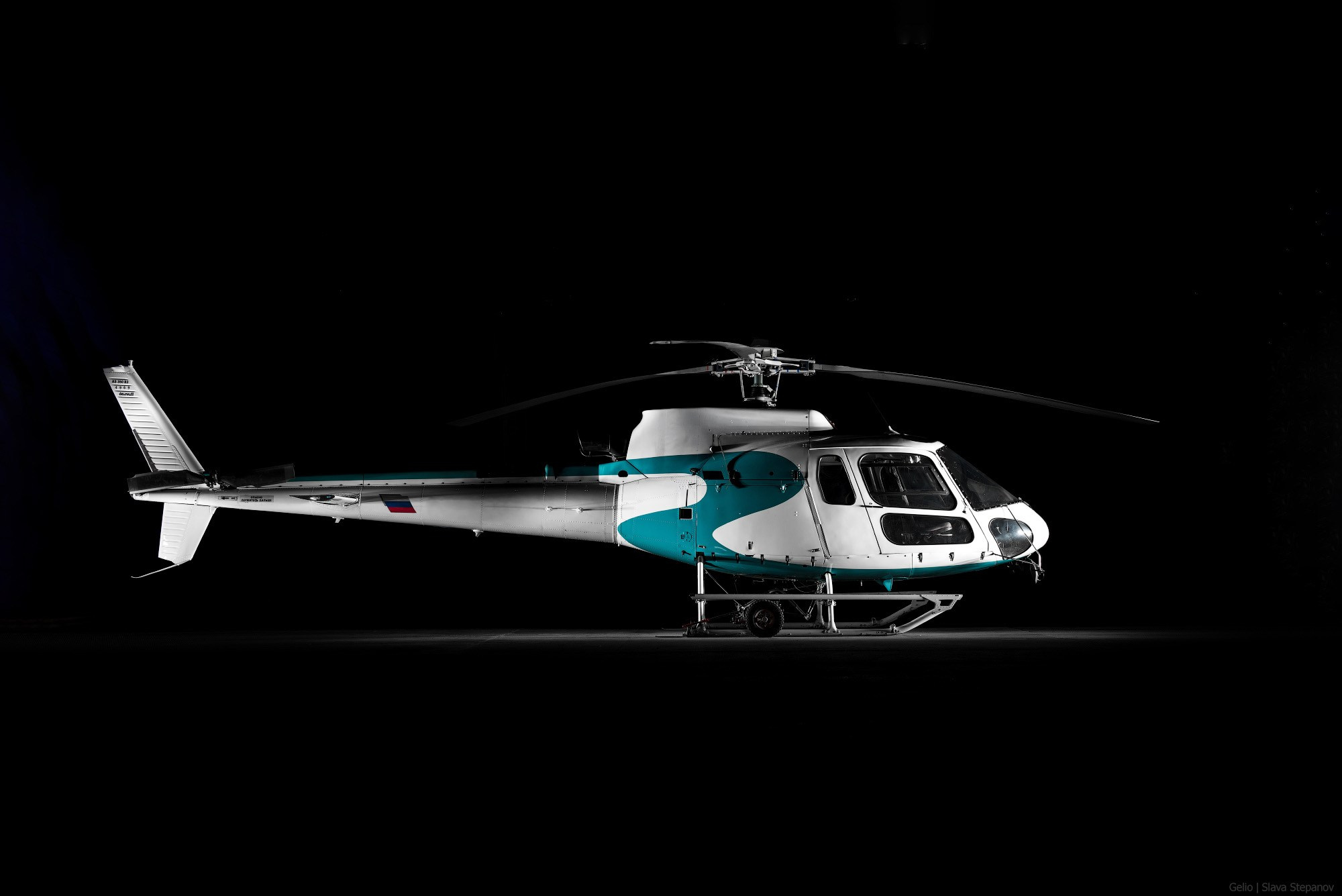 Легкий многоцелевой вертолёт Airbus Helicopters AS350 B3 в ангаре на полностью черном фоне. Боковой ракурс воздушного судна в бело-бирюзовой ливрее. Снимок Gelio.
