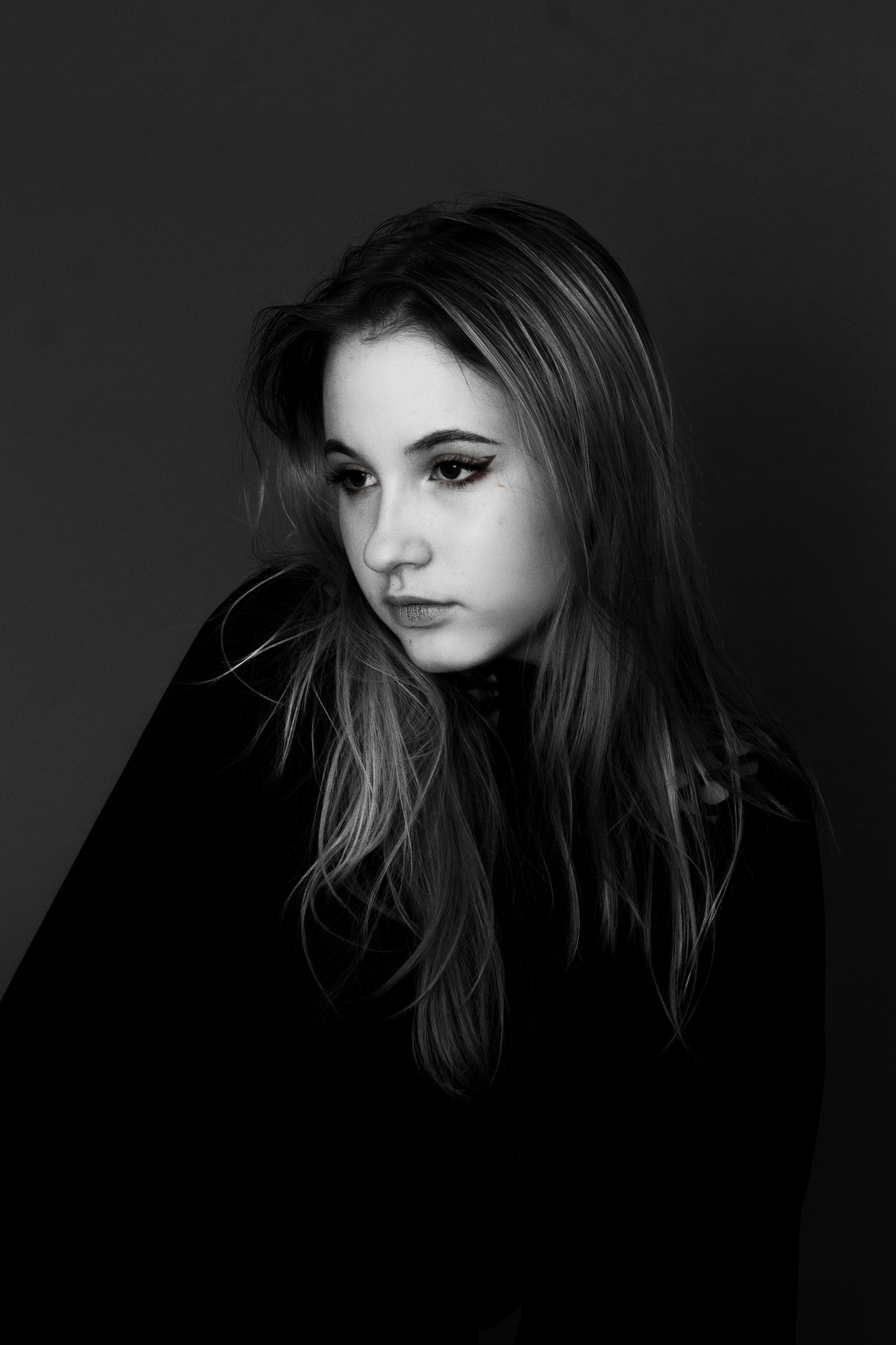 Черно-белая фотография девушки, которая для фотосессии сделала себе гранж-образ. На ней надето черное закрытое платье или блузка, глаза подчеркнуты черными стрелками, тушь густо нанесена, на губах яркий тинт.