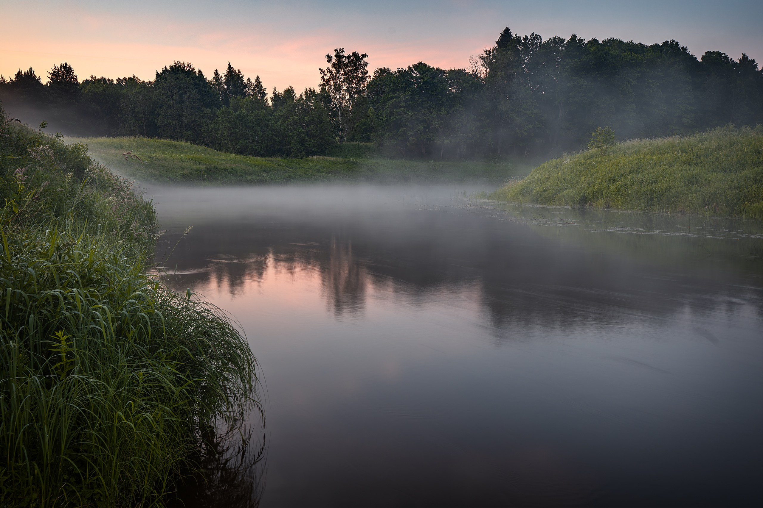Morning mist drifting above the calm Sheshupe River with trees reflected in the water at sunrise in East Prussia.