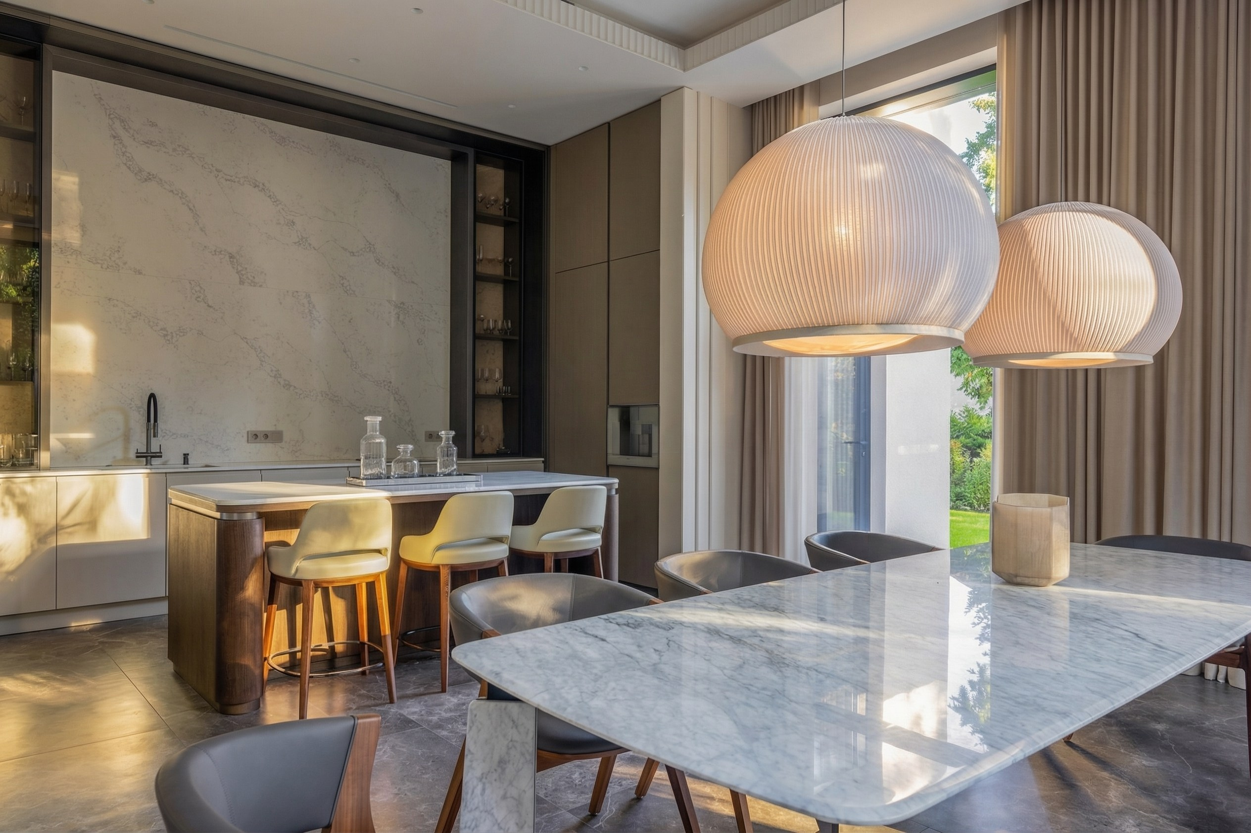 Интерьер кухни-столовой с мраморным островом, дизайнерскими барными стульями и парными светильниками-сферами, панорамный вид на сад, проект Валерии Котович в Подмосковье.