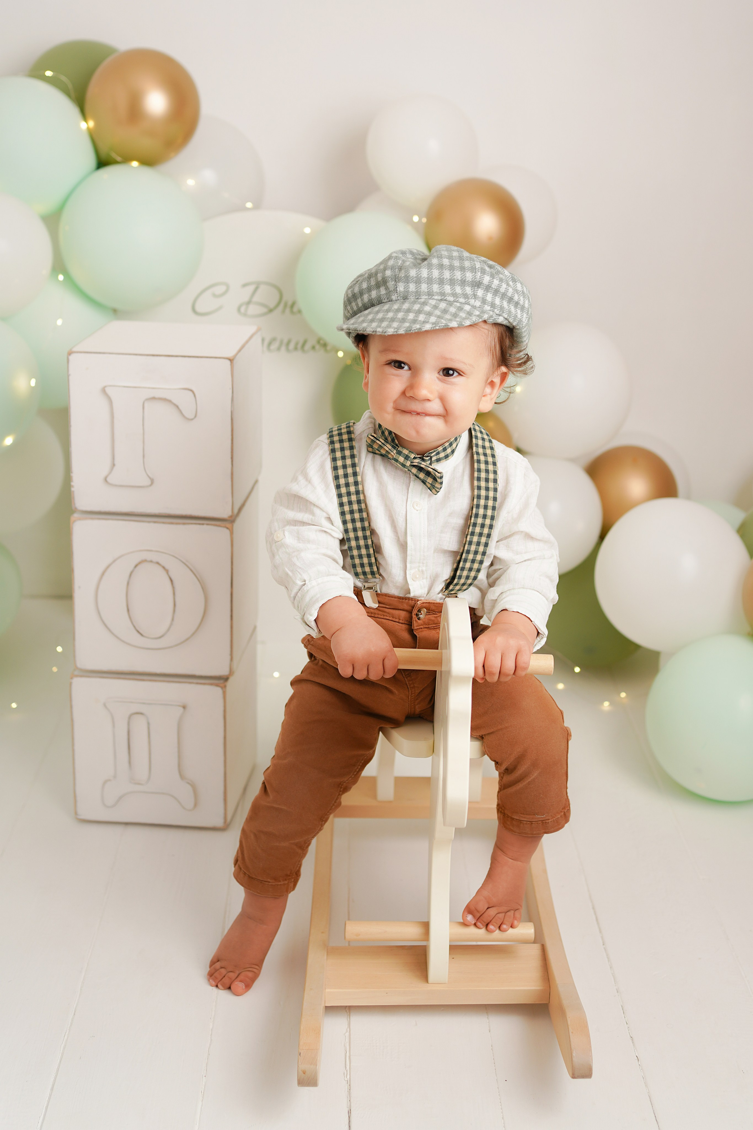 На фото малыш скачет на детской лошадке, рядом зеленые шарики, огромные кубики год. 