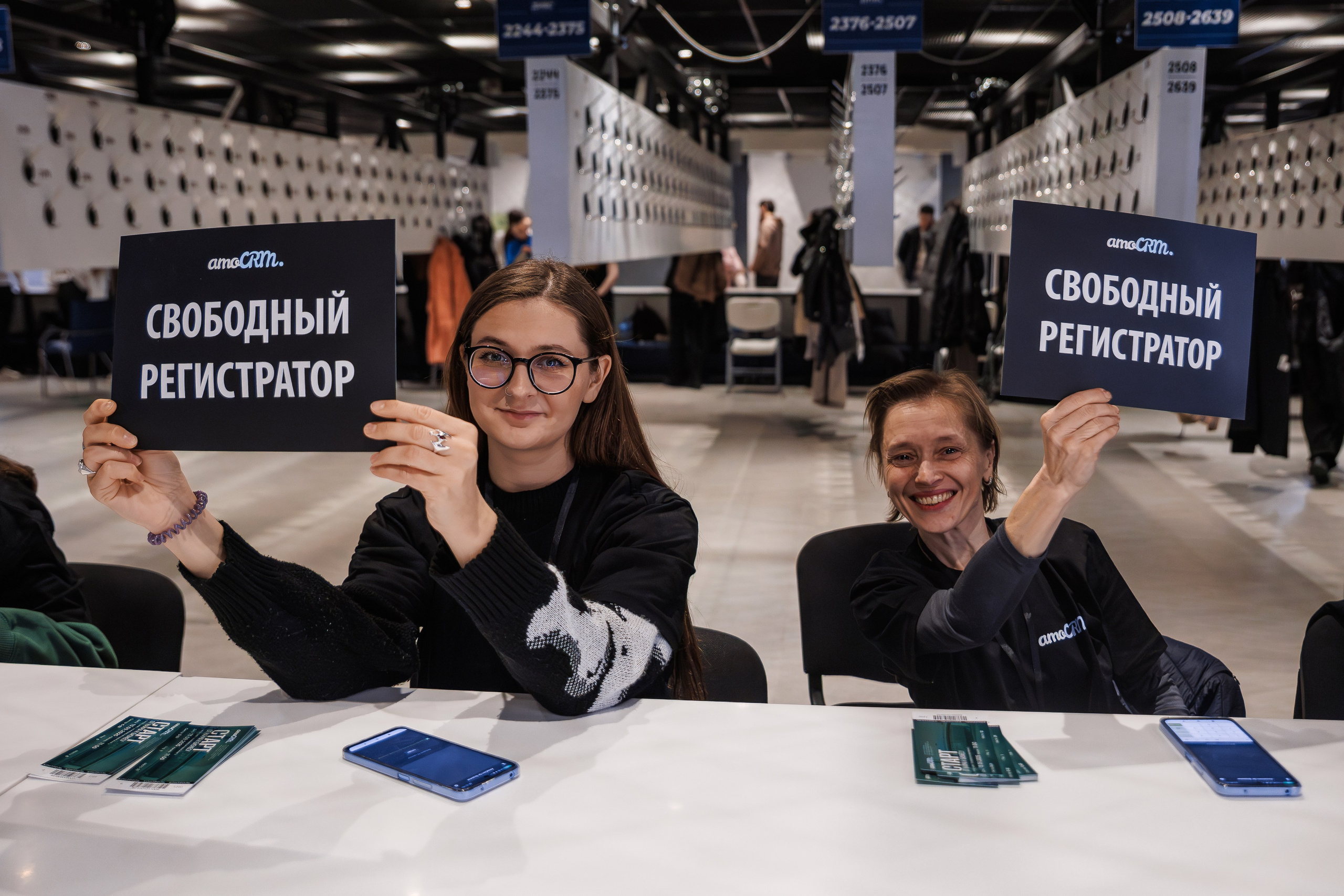 Две женщины, сидящие за регистрационной стойкой, держат таблички на русском языке с надписью «Свободная регистрация». Они улыбаются, на заднем плане — просторный современный зал с вешалками для одежды и людьми, которые ходят по залу.