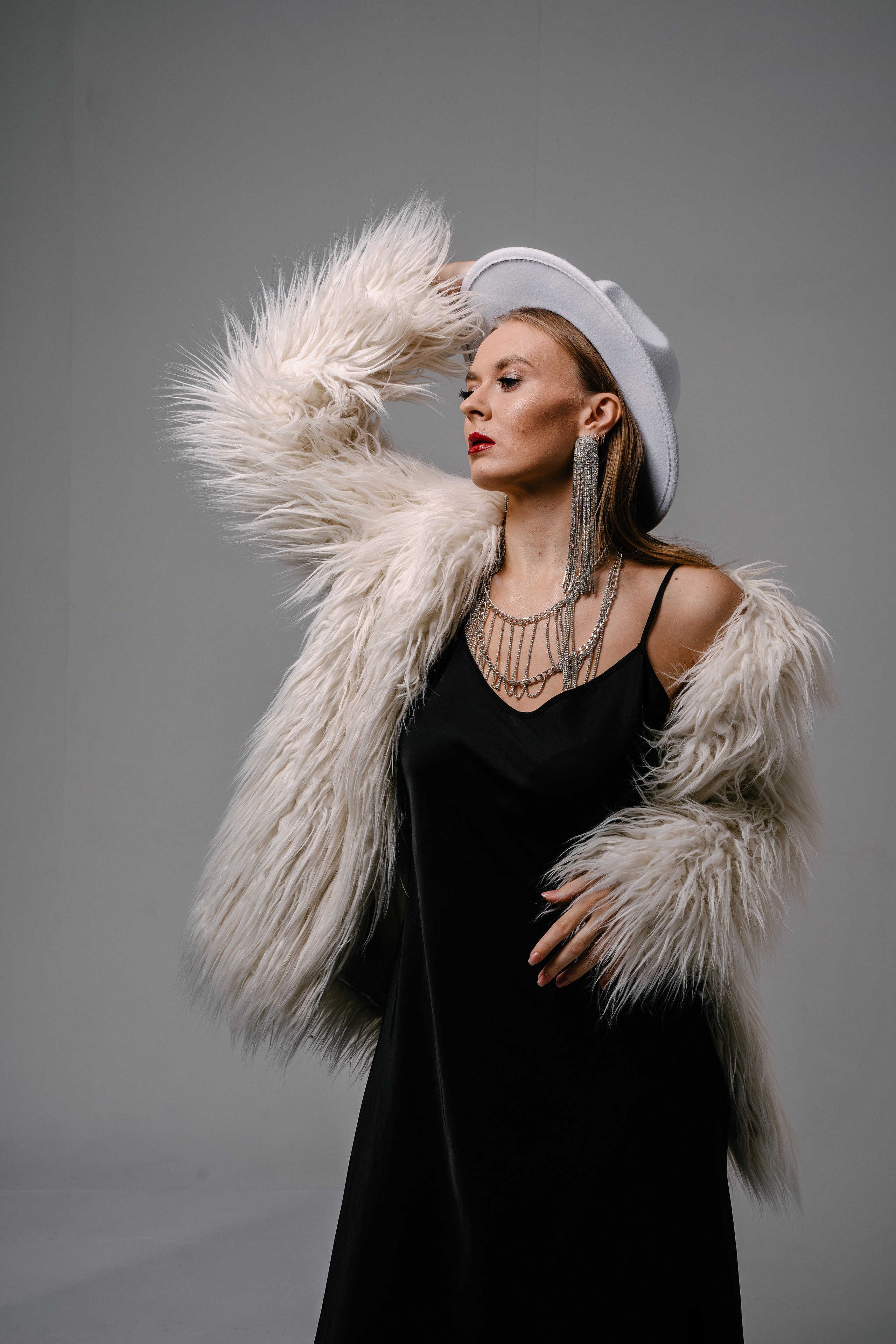 девушка в черном платье с белой шубой и в белой шляпе позирует на белом фоне