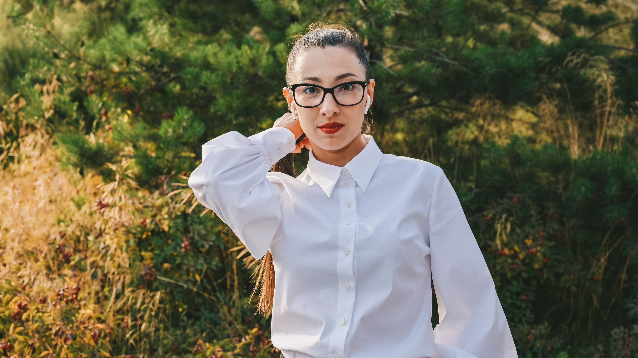 Девушка в деловом и элегантном образе с белой блузкой, черными очками и наушниками стоит на фоне живописного зеленого ландшафта. Ее длинные волосы аккуратно уложены в высокий хвост, что придает ей современный и профессиональный вид. Она одета в строгий комплект, который идеально сочетается с окружающей природной средой. Светлый фон неба и яркие краски растений создают ощущение свежести и легкости. Этот снимок демонстрирует мастерство работы с освещением и композицией, делая акцент на игре света и тени, а также на гармоничном сочетании человека и природы. Perfect for business and lifestyle photography enthusiasts!