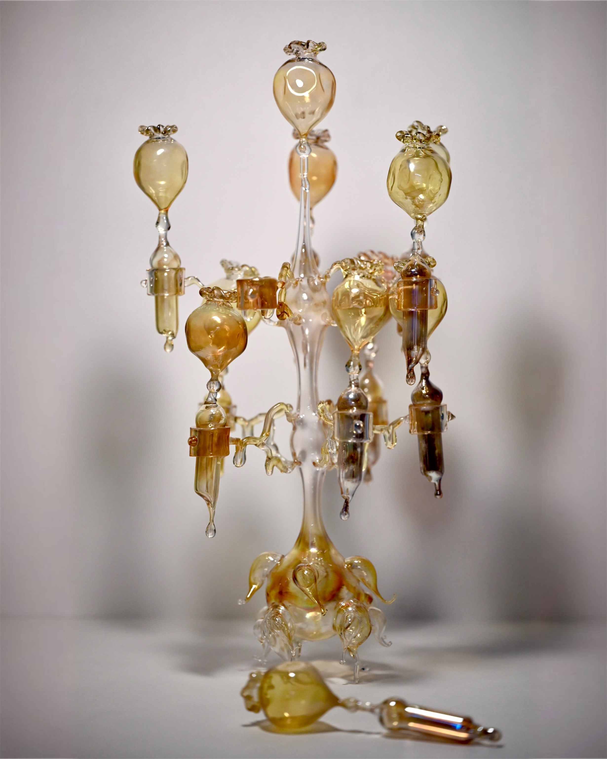 Candelabrum "Liturgy".Borosilicate glass. Lampworking. Height: 39 cm.The object refers to archaic religiosity and the image of an abandoned wooden chapel. The vertical composition is constructed as a distorted analogion: elongated forms resemble candles, deformed by time and abandonment. The yellowish smoky tone of the glass evokes dust, soot, and aged wax. The fragile, unstable structure fixes a state of loss. The candlestick functions not as a liturgical object, but as a material trace of a sacred space.